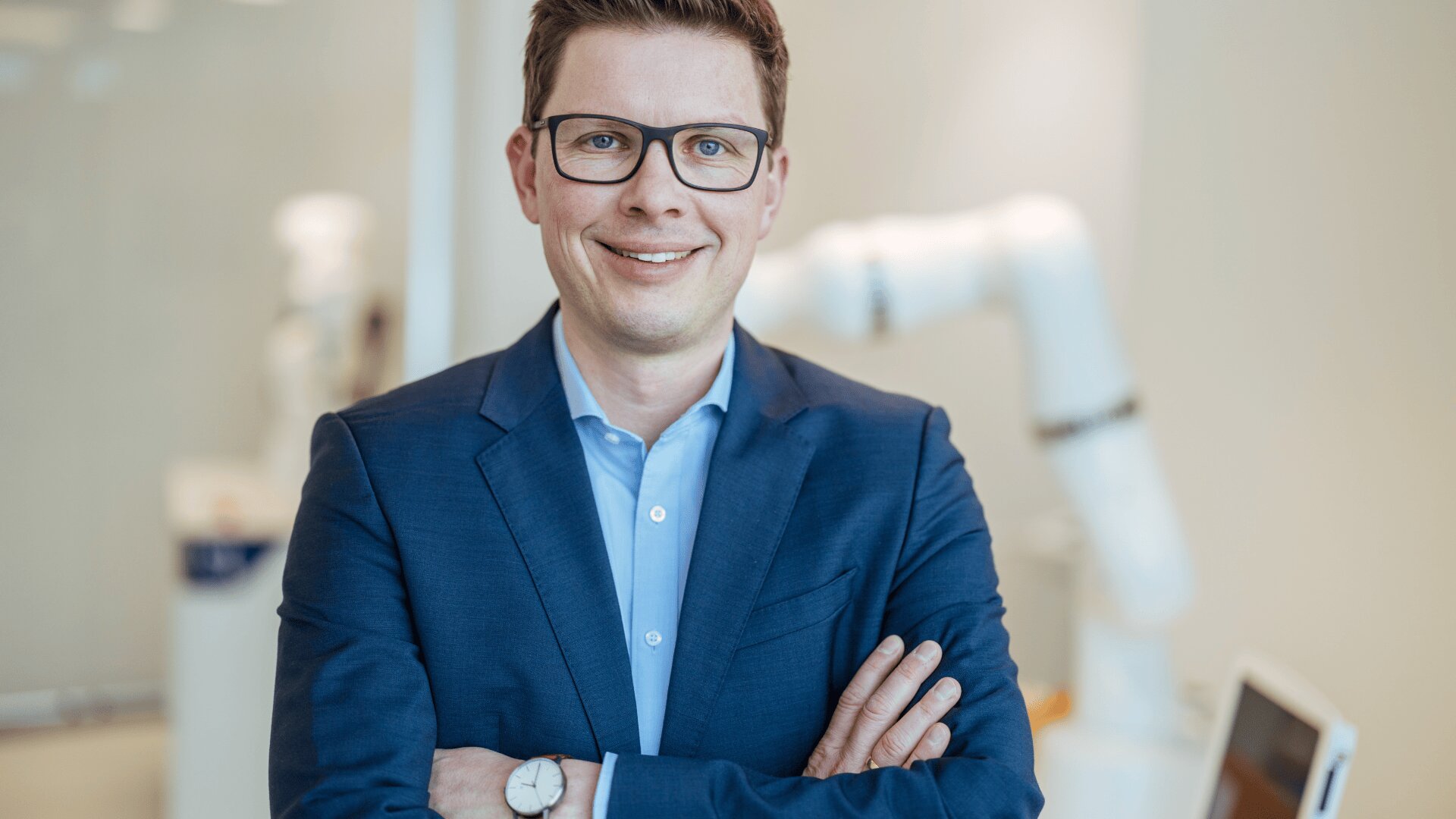 Ein Mann in einem blauen Anzug und mit Brille steht mit verschränkten Armen da und lächelt selbstbewusst. Der Hintergrund ist ein heller, moderner Raum mit verschwommenen weißen Roboterarmen und Geräten.