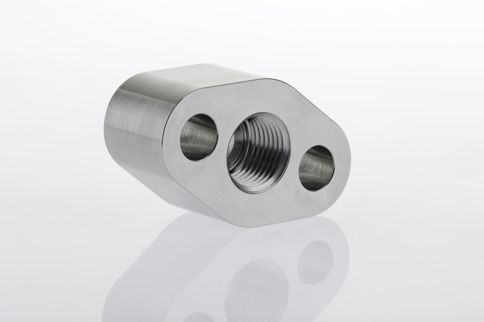 A machined metal component with a cylindrical body and three circular holes, one threaded and two smooth, placed on a reflective white surface.