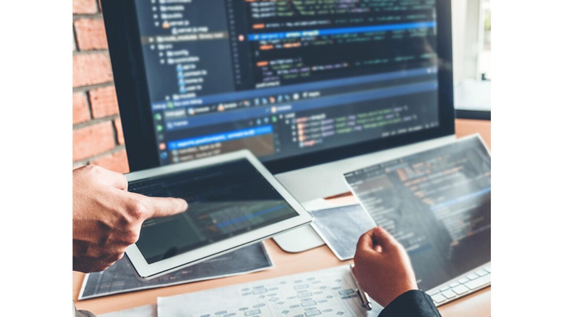 Two people work at a desk with a computer displaying code. One person points at a tablet, while the other holds a transparent sheet with printed code. Papers with diagrams are scattered on the desk.
