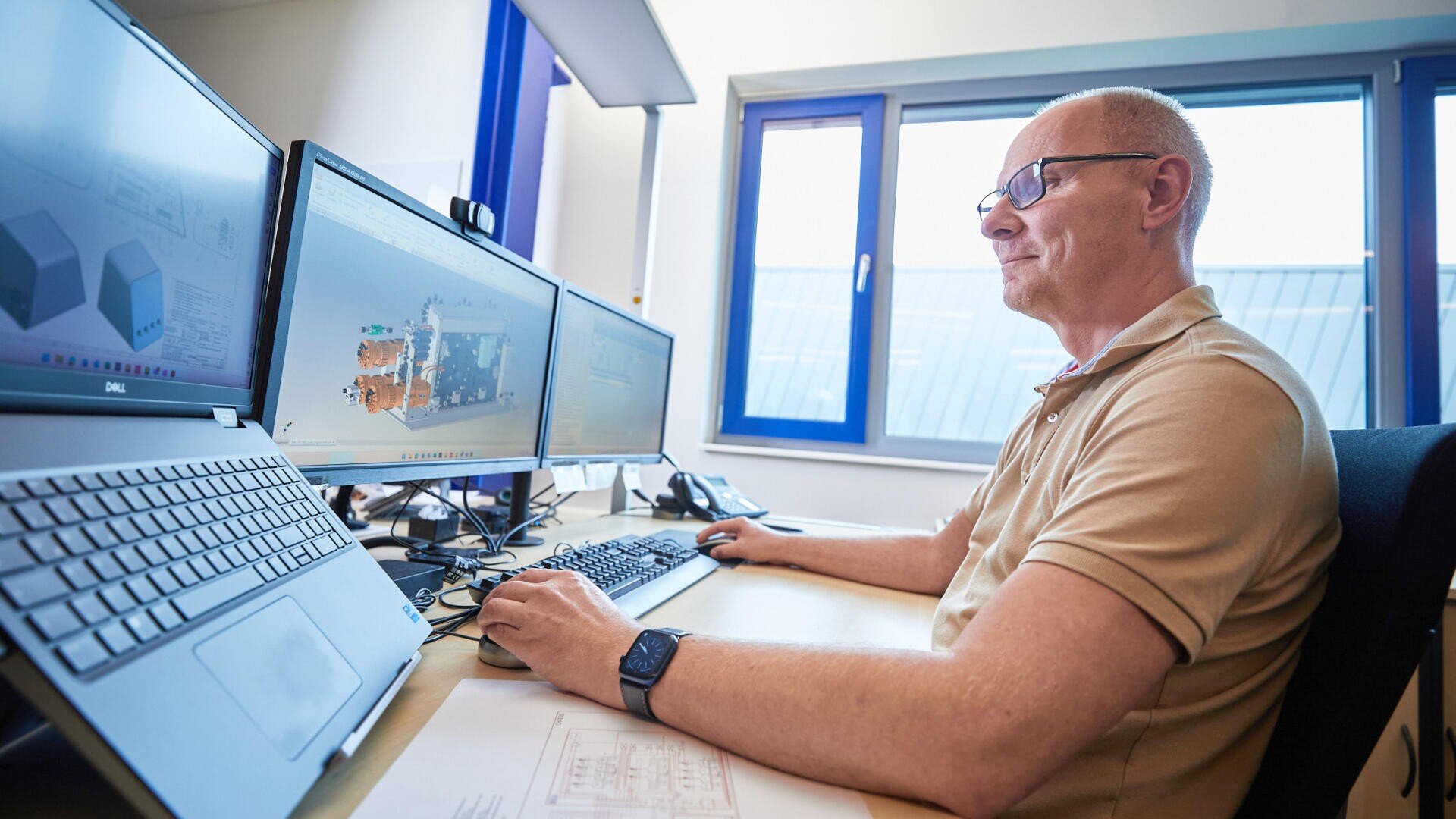 Ein Mann mit Brille arbeitet an einem Schreibtisch mit drei Computermonitoren, auf denen technische oder ingenieurtechnische Entwürfe angezeigt werden, in einem hellen Büro mit Fenster.
