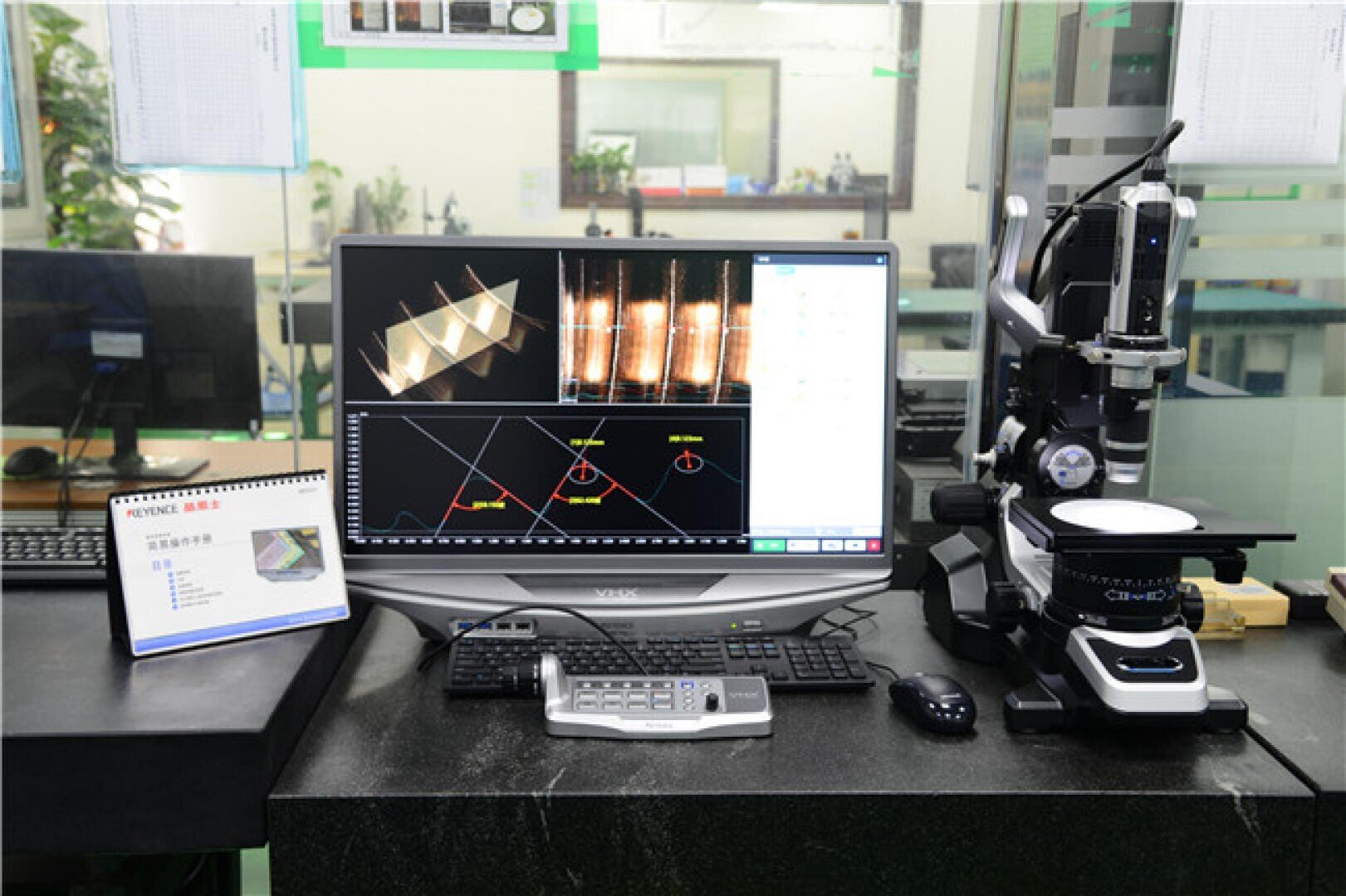 A laboratory workstation with a computer displaying scientific graphs and images, a sophisticated microscope to the right, a control pad, mouse, keyboard, and an open instruction manual on the left.