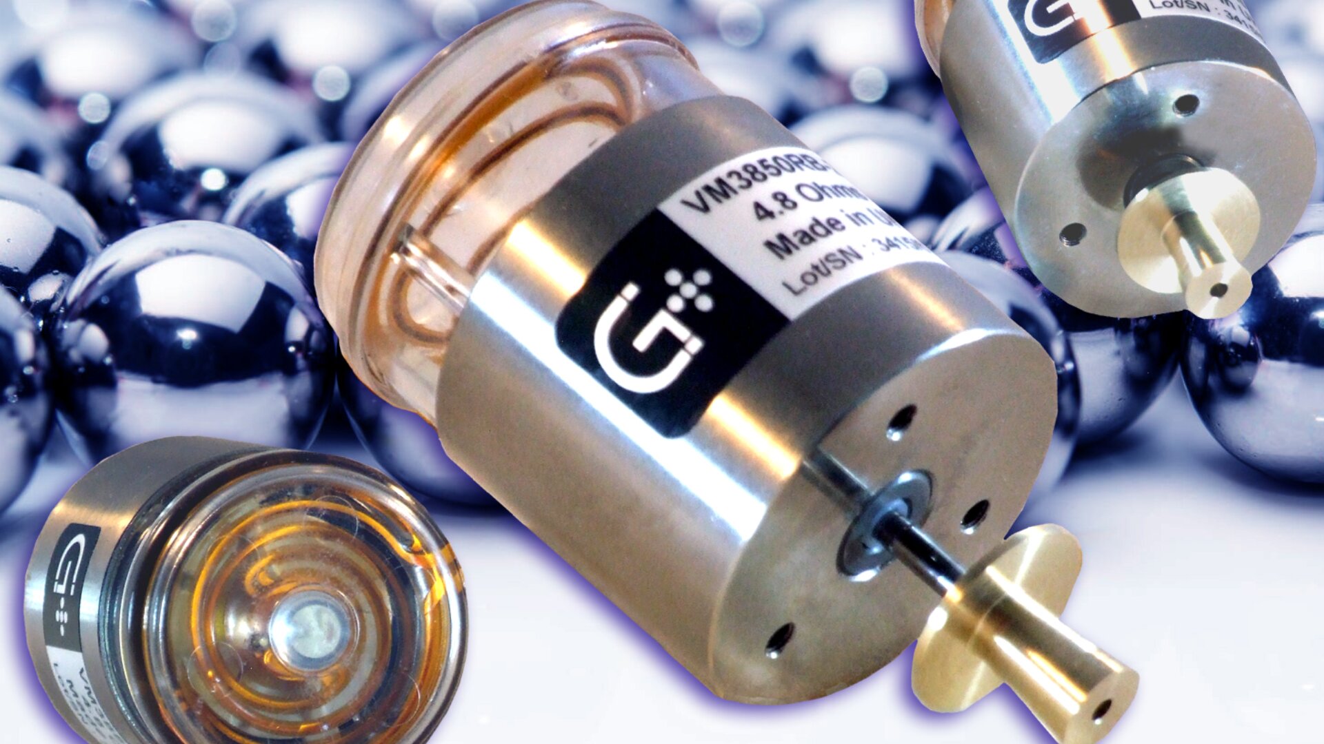 Nahaufnahme von drei kleinen Metallmotoren mit Aufklebern, die vor einem Hintergrund aus glänzenden Metallkugeln stehen. Die Motoren sind zylindrisch und bestehen aus metallischen und transparenten Teilen, wobei einer von unten gezeigt wird.