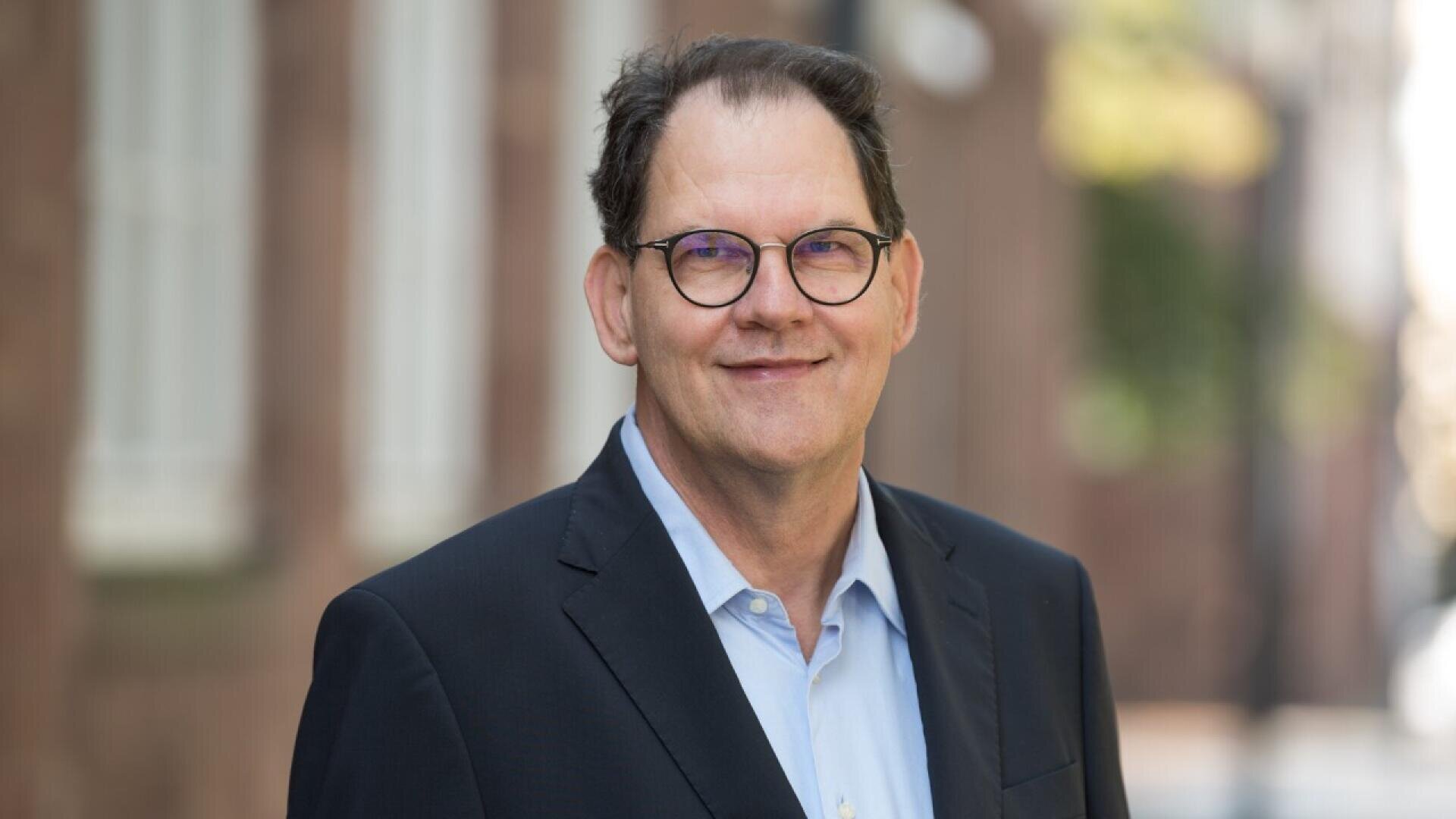 A middle-aged man with short dark hair wearing glasses, a light blue shirt, and a dark suit jacket smiles outdoors with blurred buildings in the background.