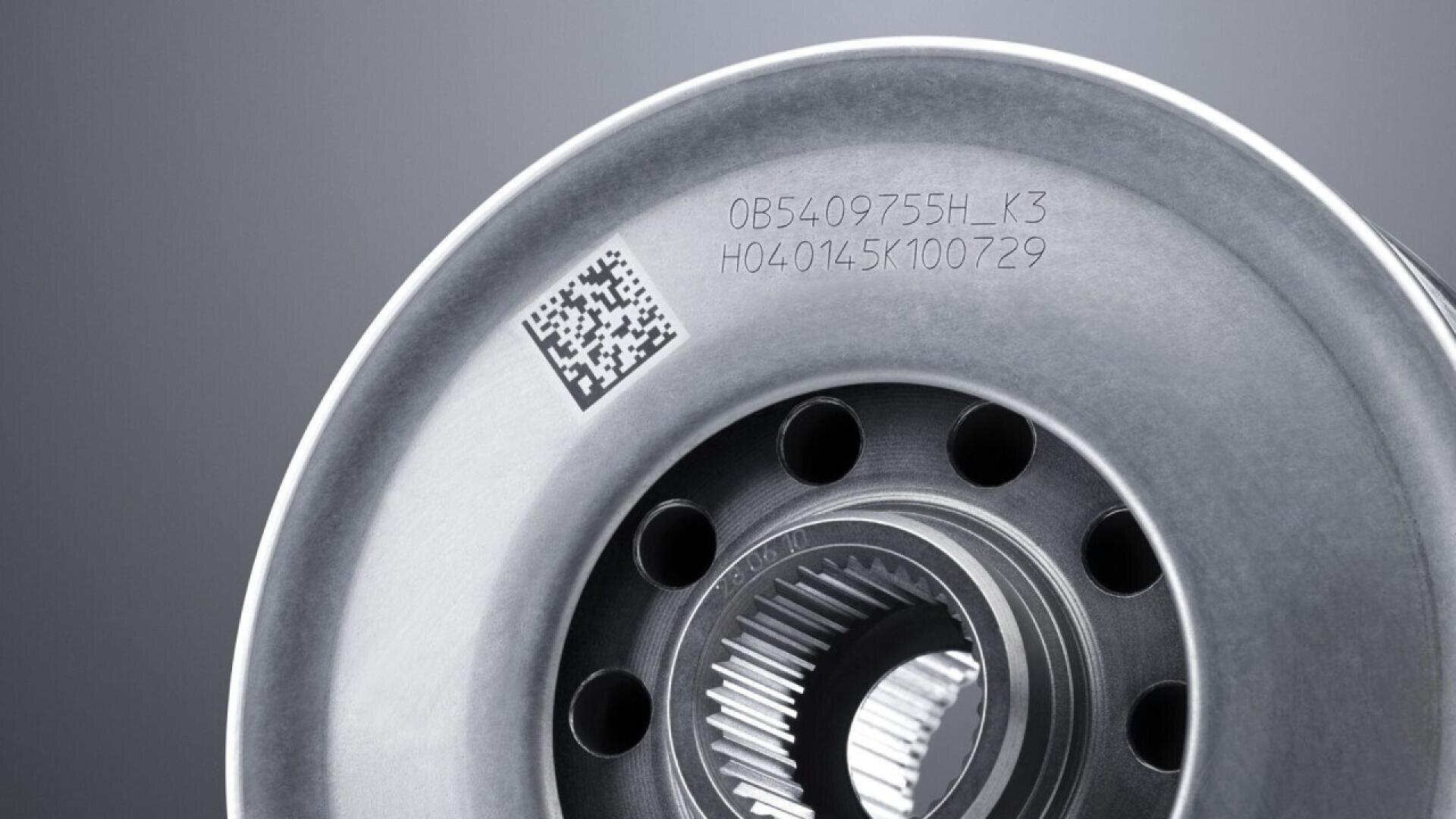 A close-up of a metallic machine part with several holes and engraved alphanumeric codes, along with a small QR code on its smooth, circular surface. The background is blurred.