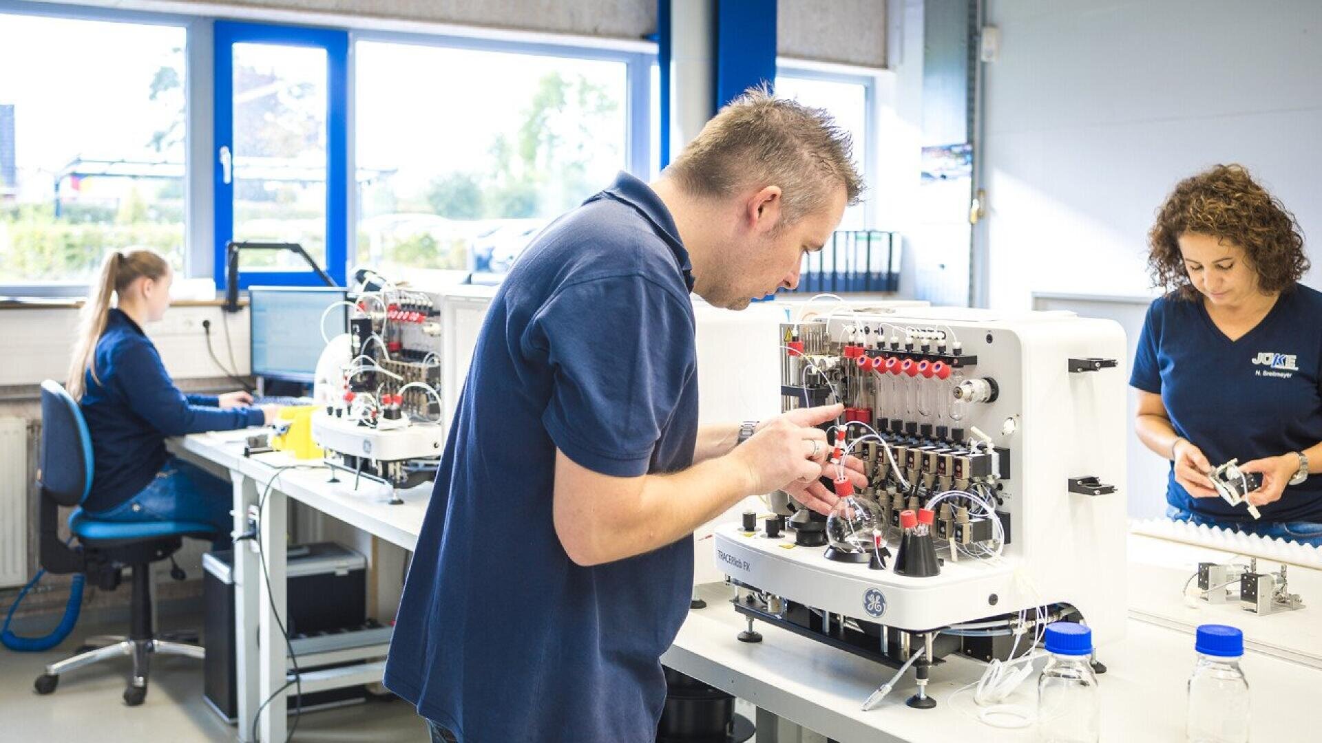Three people work in a bright laboratory; one man focuses on equipment assembly, a woman assembles parts at another station, and a third person works at a computer near a window. Lab devices and bottles are on the tables.