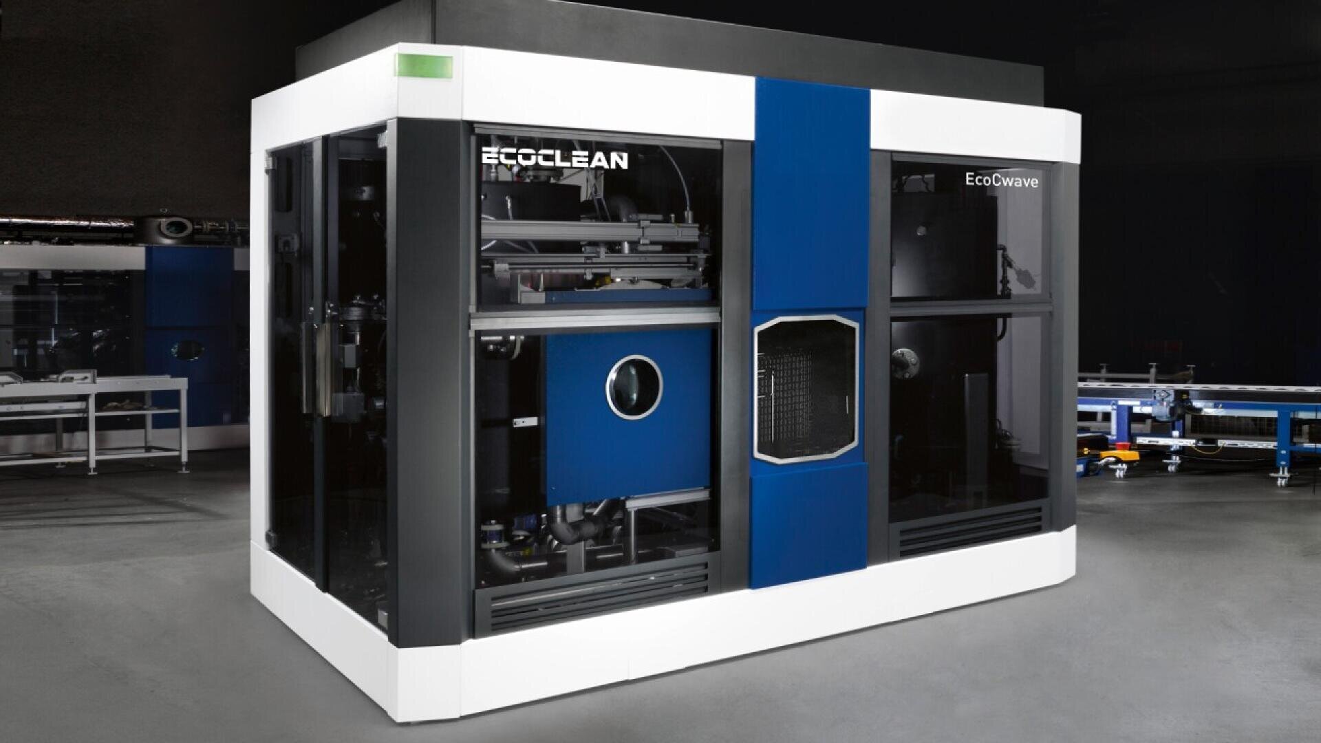 Large industrial machine with a white and blue exterior, labeled Ecoclean and EcoCwave, featuring glass panels showing internal mechanical components, situated in a clean, modern facility.