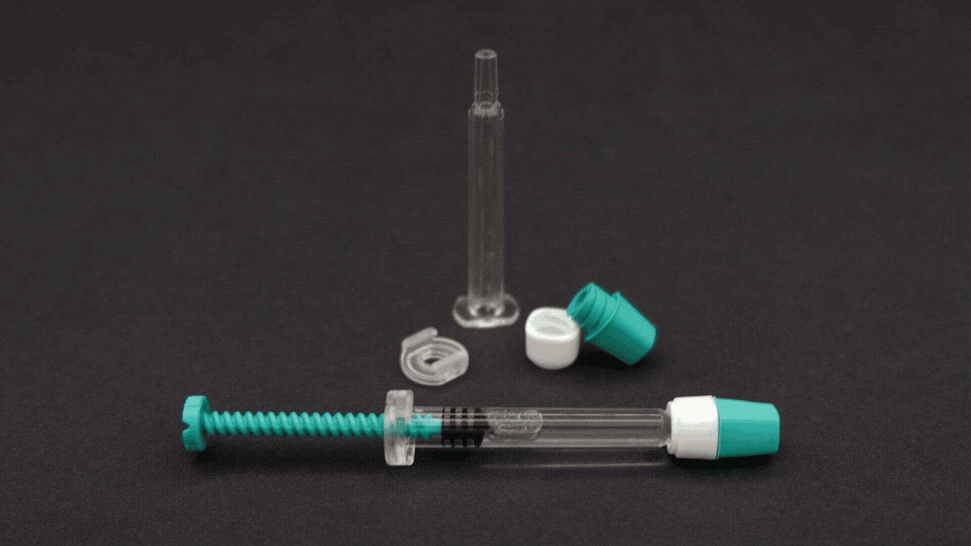 Eine durchsichtige Plastikspritze mit grünem Kolben, grüner und weißer Kappe und einer durchsichtigen Schutzhülle ist auf einer schwarzen Fläche ausgestellt. Die Komponenten sind getrennt und liegen neben der Spritze.