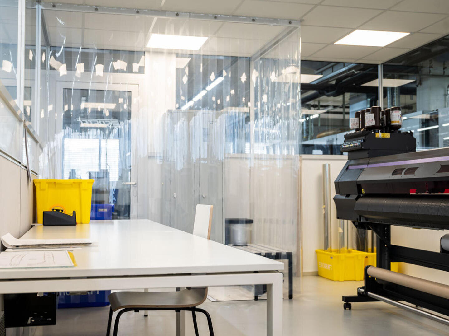 Ein sauberer, moderner Arbeitsplatz mit weißem Schreibtisch, Stuhl und Papieren neben einem großen Drucker. Gelbe Mülleimer und transparente Plastikvorhänge trennen den Bereich ab. Der Raum ist hell beleuchtet und minimalistisch gestaltet.