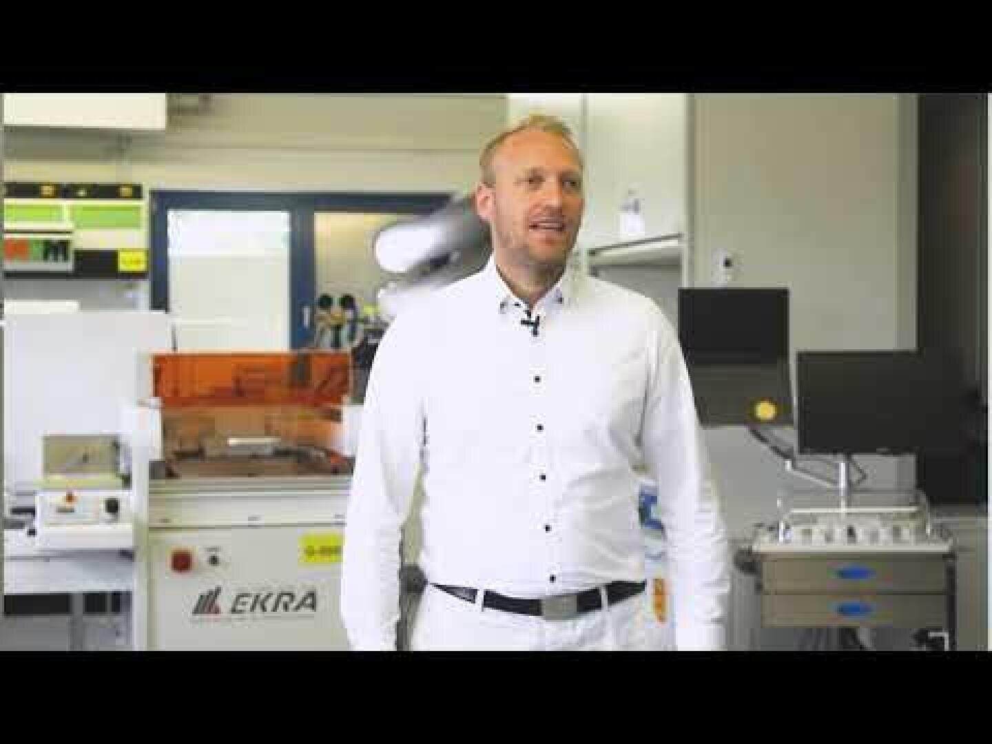 Ein Mann in einem weißen Hemd steht in einem modernen Labor oder einer Produktionsstätte, in der verschiedene Geräte und Maschinen, darunter eine mit der Aufschrift EKRA, im Hintergrund zu sehen sind.