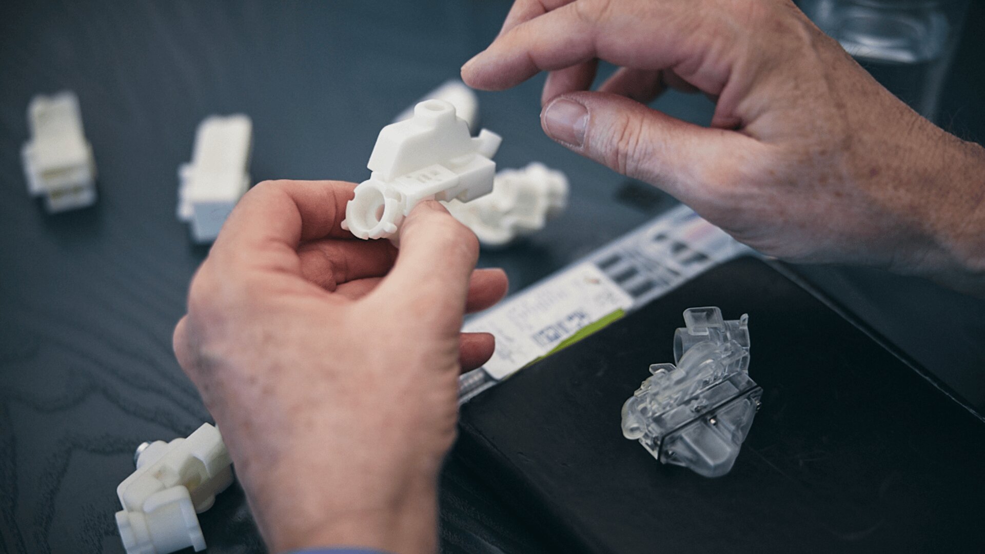 Nahaufnahme von Händen, die ein weißes 3D-gedrucktes mechanisches Teil halten und untersuchen, mit weiteren 3D-gedruckten Objekten und Werkzeugen auf einem dunklen Tisch im Hintergrund.