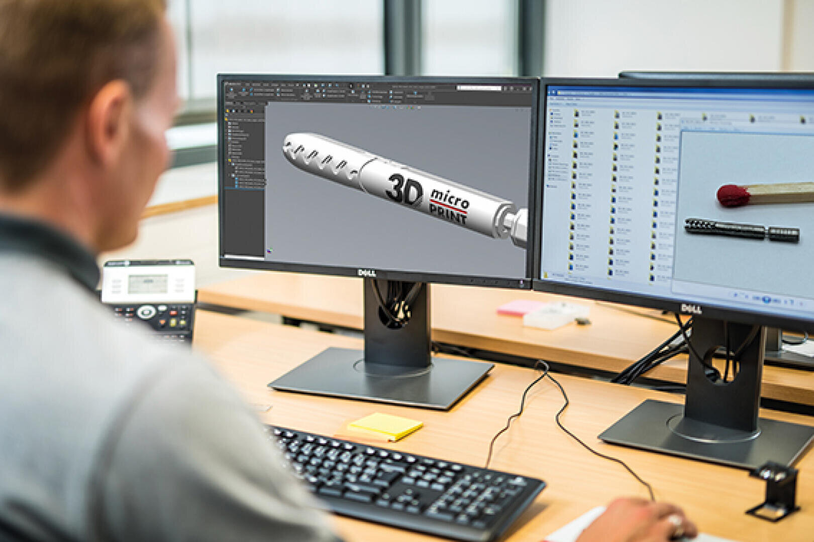 A person works at a desk with two monitors; one screen shows a 3D model of a metallic object labeled 3D micro PRINT, and the other displays a photo of the same object next to a matchstick for scale.