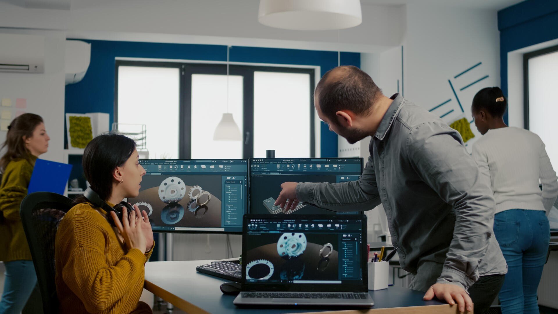 Zwei Kollegen diskutieren in einem Büro über 3D-Konstruktionen von mechanischen Getrieben, die auf Computermonitoren angezeigt werden, während drei weitere Personen im Hintergrund arbeiten.