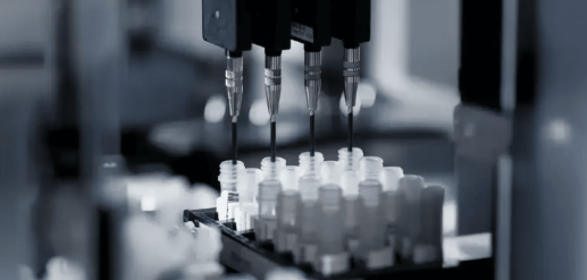 Close-up of an automated laboratory machine dispensing liquid into rows of small transparent sample tubes using multiple pipettes. The background is blurred, highlighting the precision equipment.