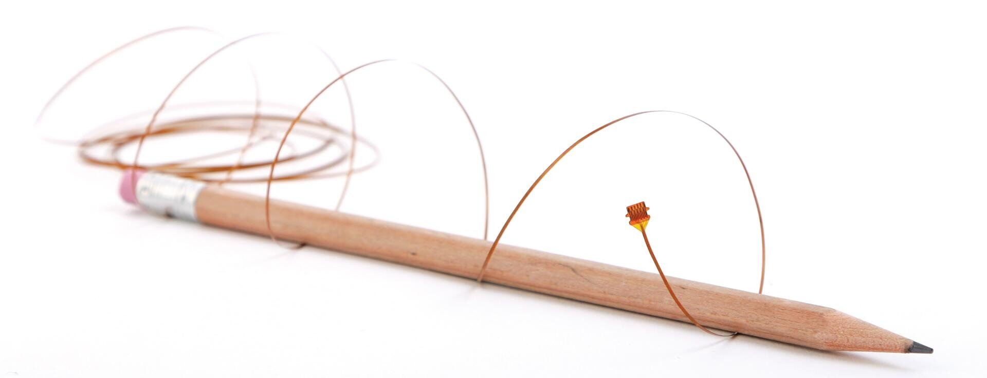 A wooden pencil lies horizontally on a white surface with a thin copper wire coiled loosely around it. A small orange electronic component is attached to the wire near the pencil tip.