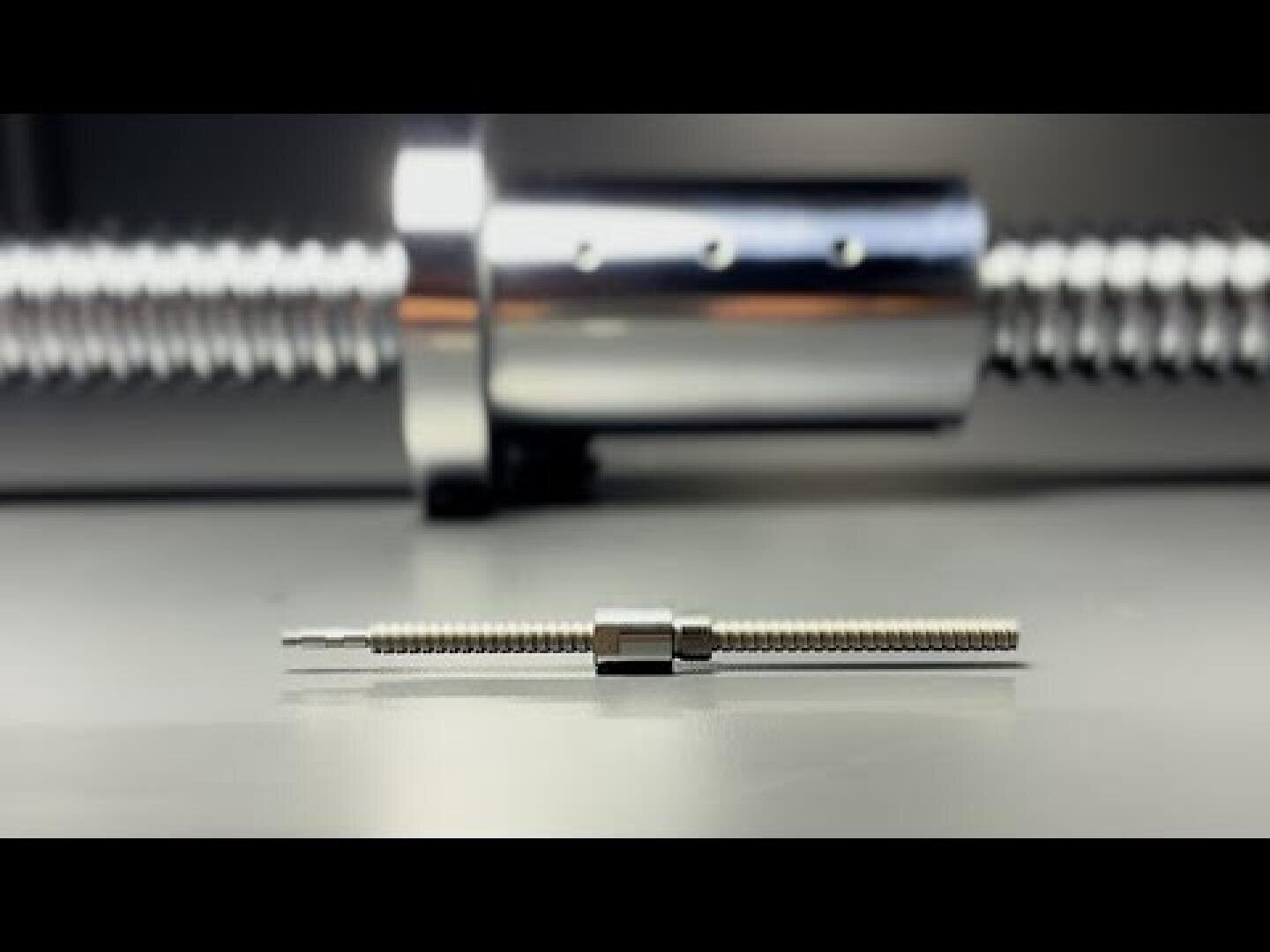 A close-up of a small metal spindle with matching nut on a gray surface, with a larger, blurred threaded spindle and nut in the background.