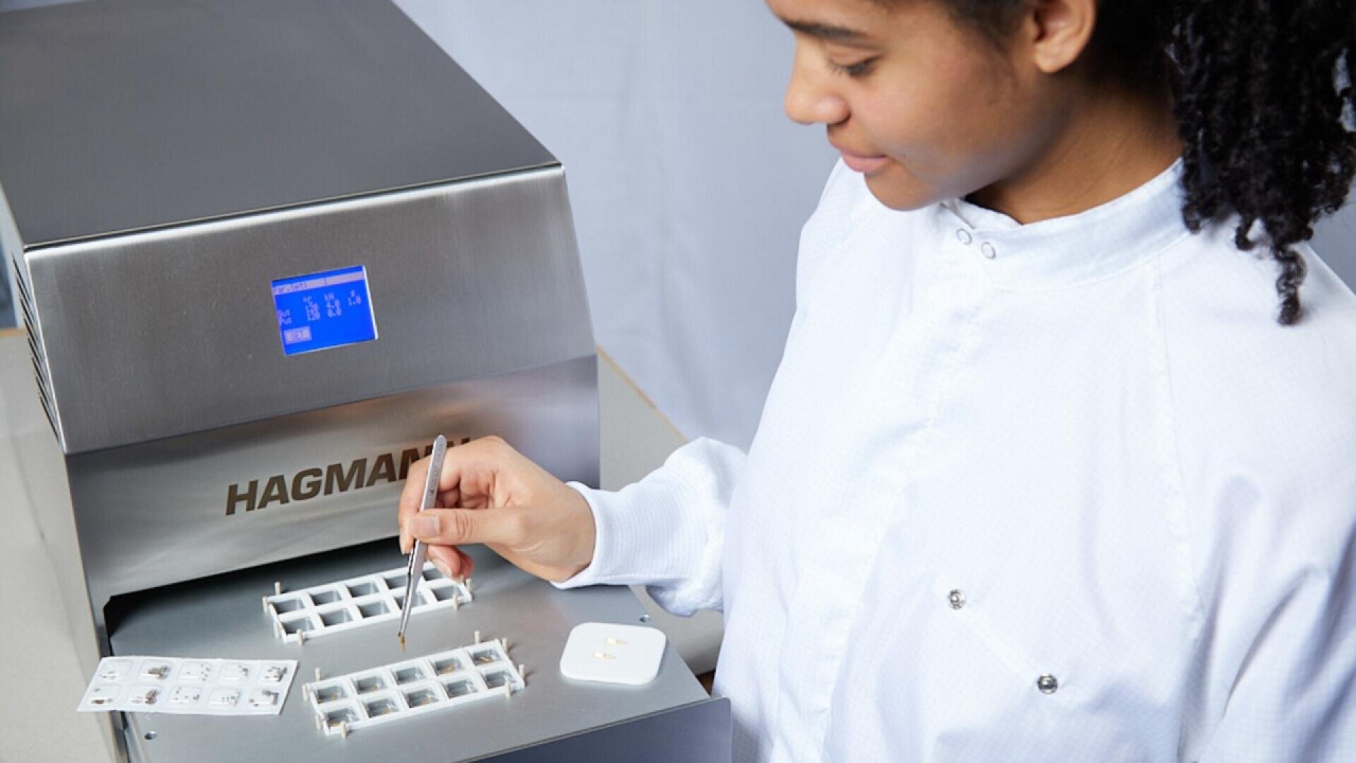 A scientist in a white lab coat uses tweezers to place samples into a metal laboratory machine labeled HAGMAN with a small digital display screen.