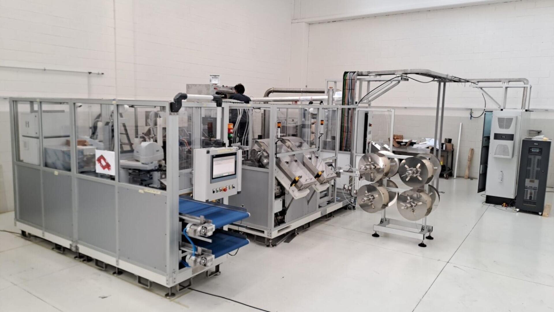 Industrial machinery with conveyor belts and metal rollers in a clean, white factory setting. The equipment appears to be automated, with control panels and various components enclosed in glass.