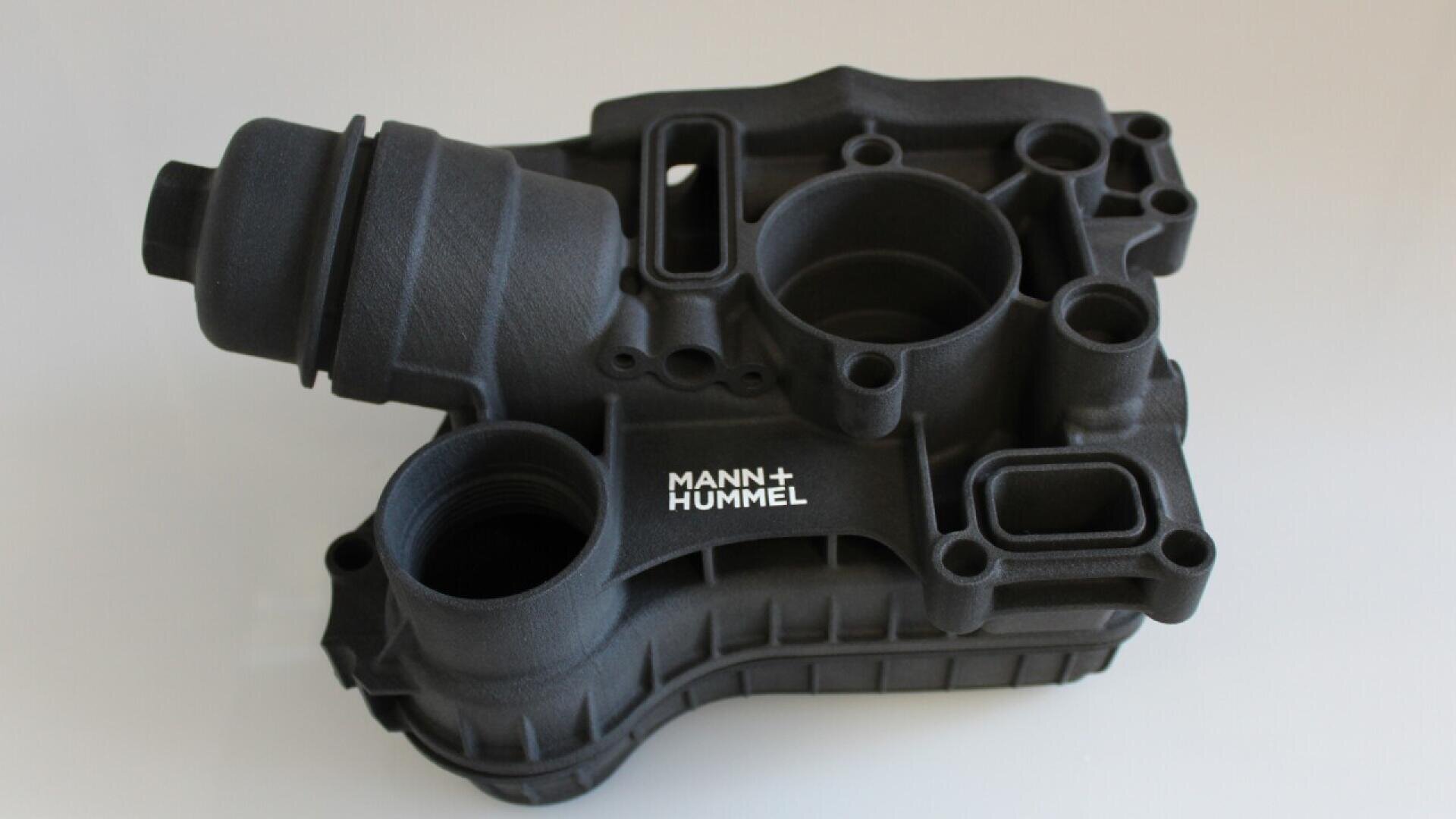 A black, intricately shaped automotive engine part labeled MANN+HUMMEL sits on a plain, light background. The component features multiple round openings and structural details.