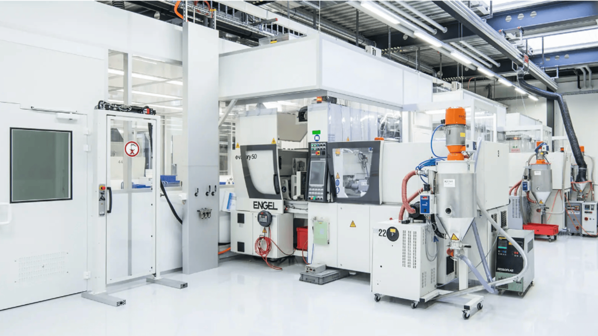 Eine saubere, moderne Fabrik mit weißen Böden und Wänden. In ihr befinden sich mehrere große automatisierte Maschinen und Anlagen für Fertigungs- oder Produktionsprozesse. Der Bereich ist hell mit Deckenleuchten beleuchtet.