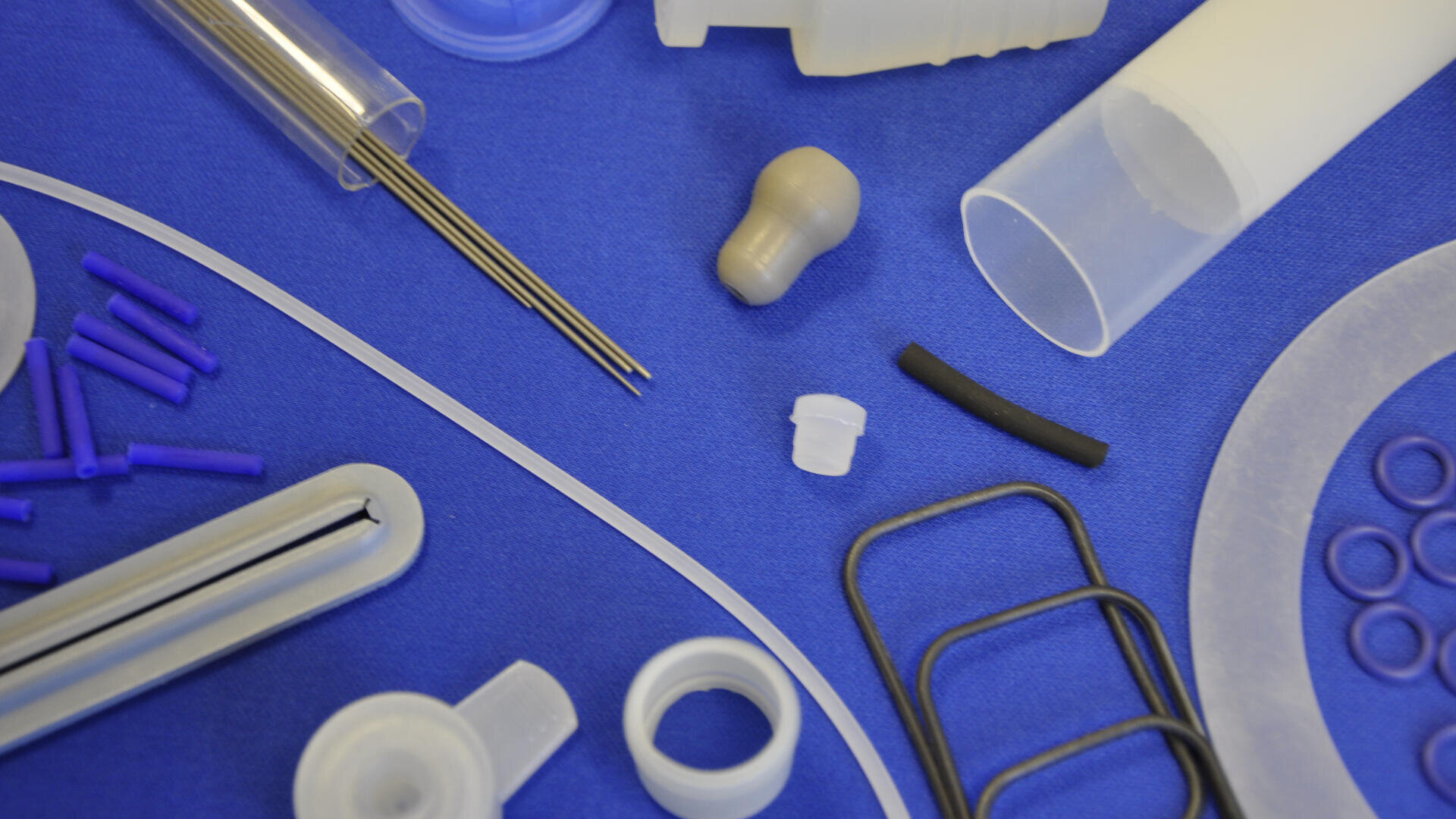 A variety of plastic and metal mechanical components, including tubes, rings, rods, and fittings, are arranged on a blue surface. The items are of different shapes, sizes, and colors, mainly white, black, and purple.