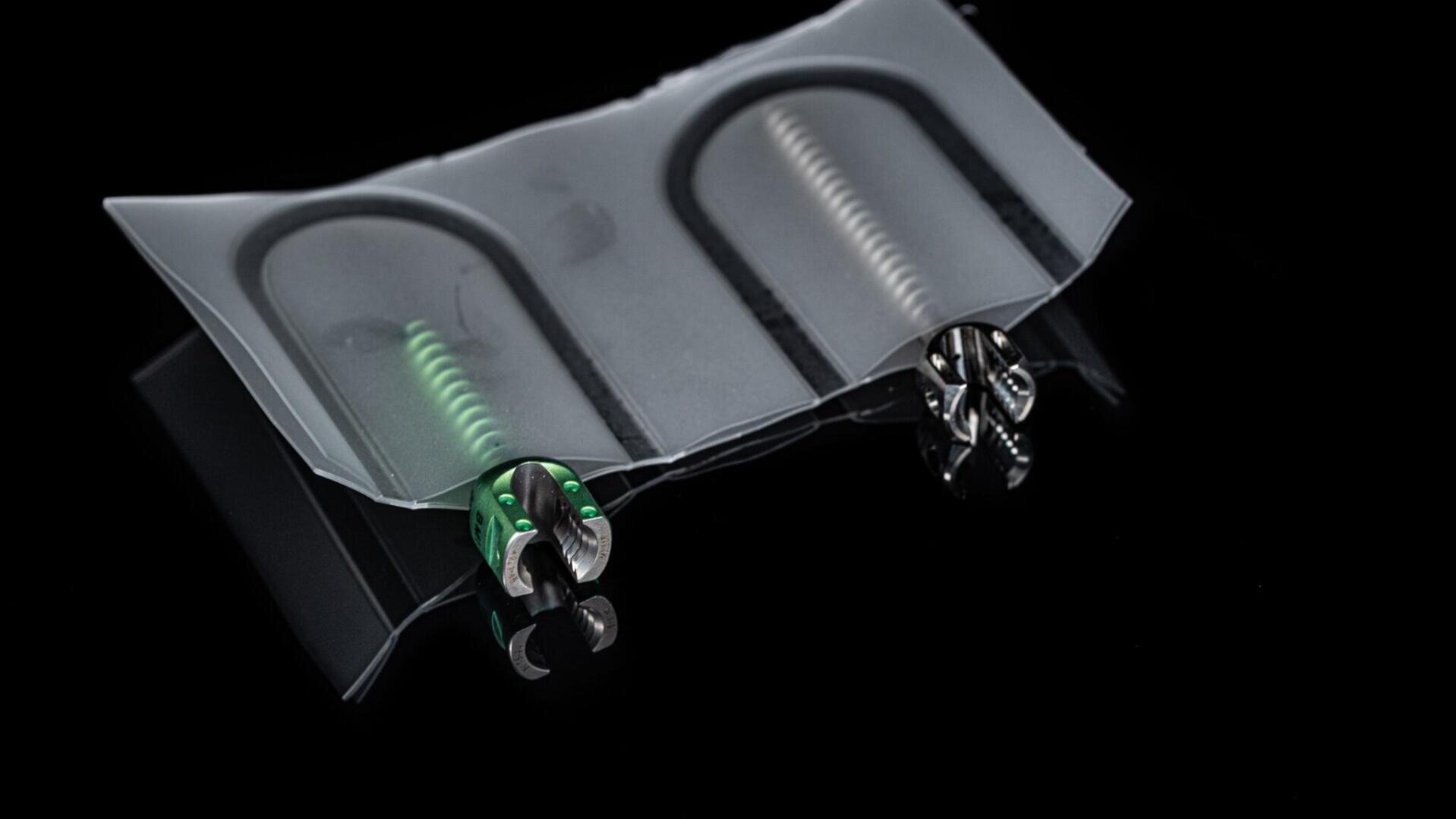 Two dental endodontic files, one with a green handle and one with a silver handle, are partially enclosed in a transparent plastic sterile pouch against a black background.