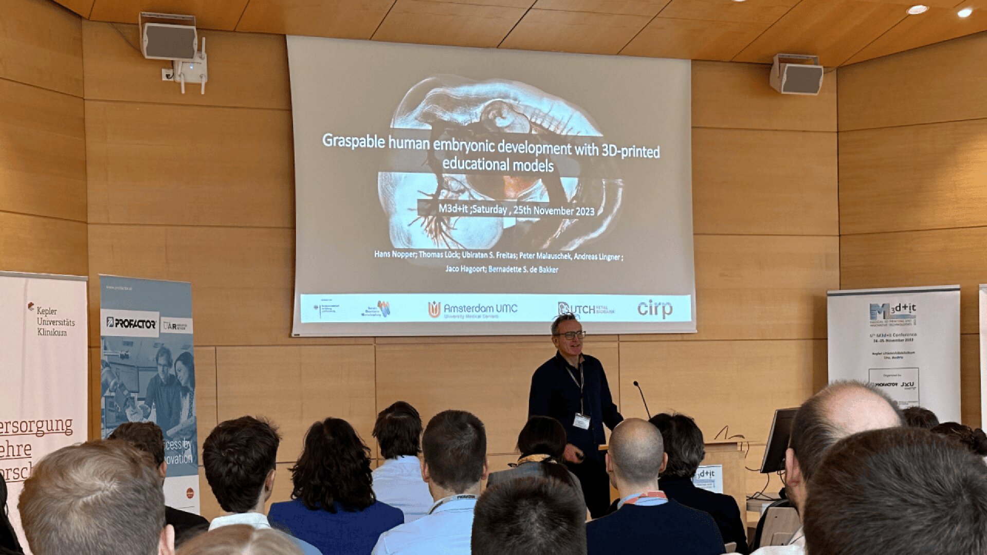 A speaker presents to an audience in a lecture hall. The projected slide behind him reads, “Graspable human embryonic development with 3D-printed educational models,” with event and contributor details. Banners and logos are visible.