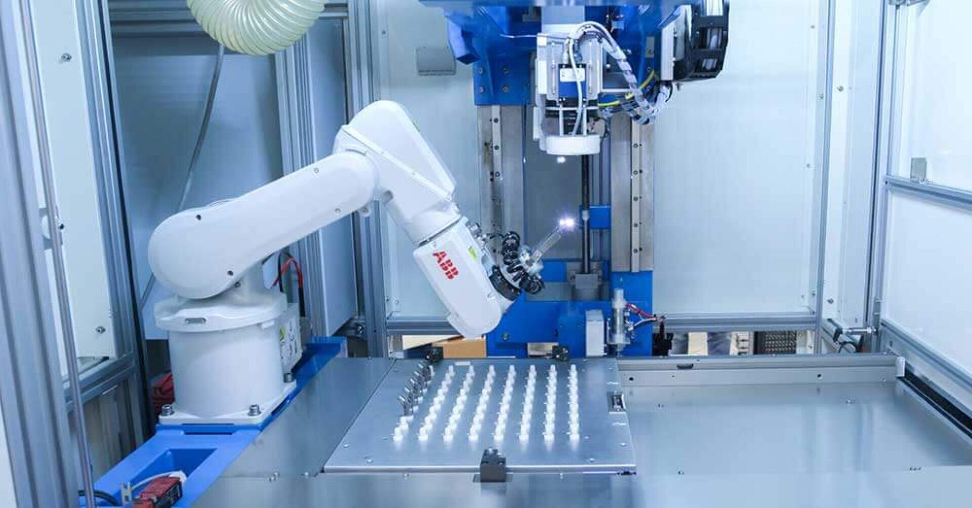 A robotic arm works in a closed industrial workplace, precisely handling small white objects arranged in a grid on a metal surface, while automated machinery and cables are seen in the background.