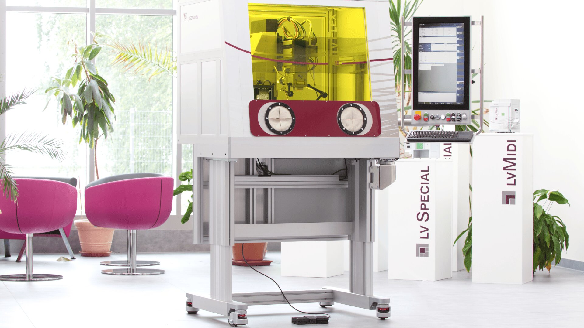 Eine Labormaschine mit gelber Sicherheitsabdeckung, zwei Handschuhöffnungen und einem Touchscreen-Bedienfeld steht in einem hellen Raum mit modernen rosa Stühlen und grünen Pflanzen.