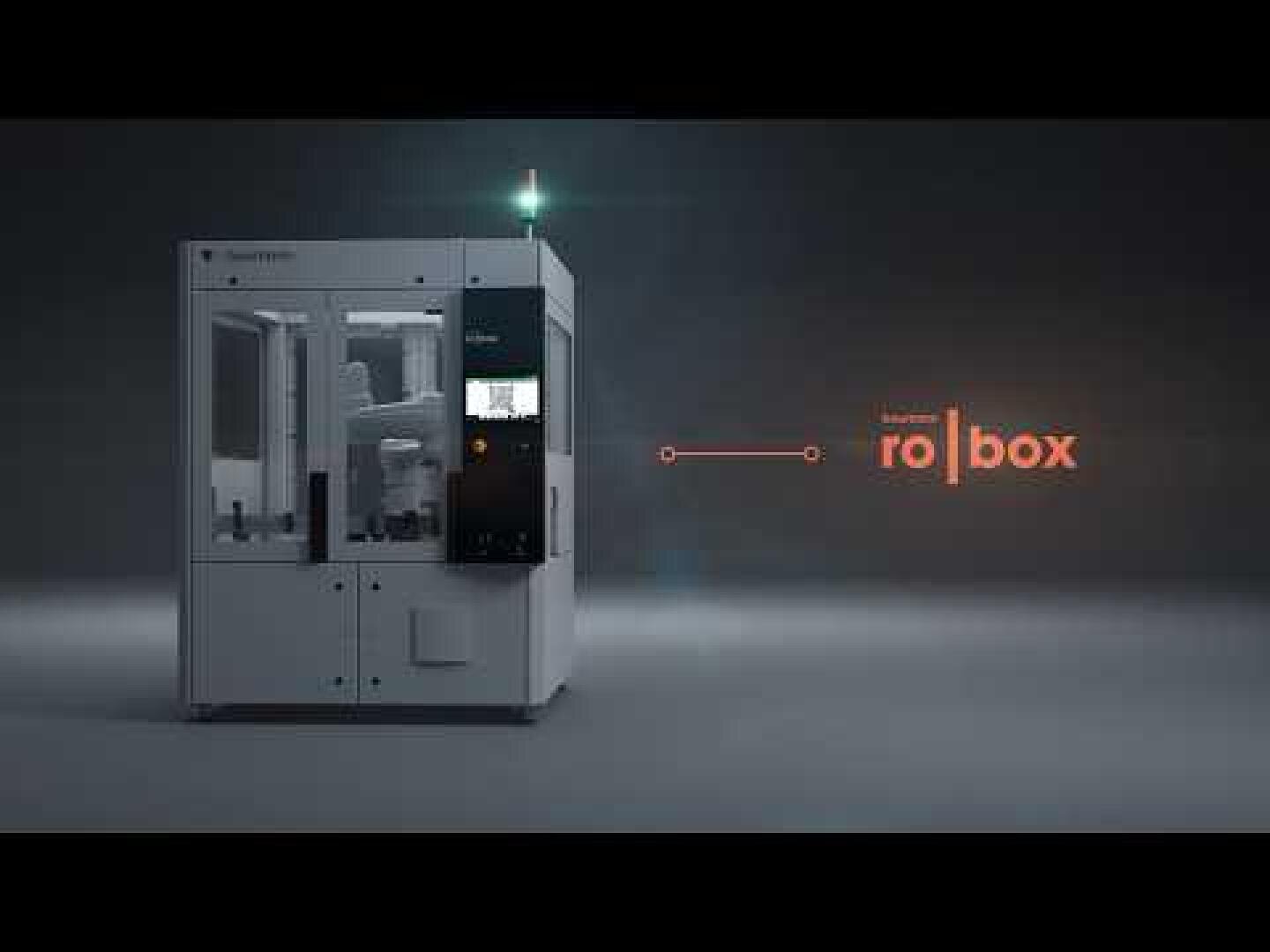 A large, white industrial machine with a glass front and a small screen stands in a dark room. An orange line connects the machine with the glowing Krones ro|box lettering on the right-hand side of the picture.