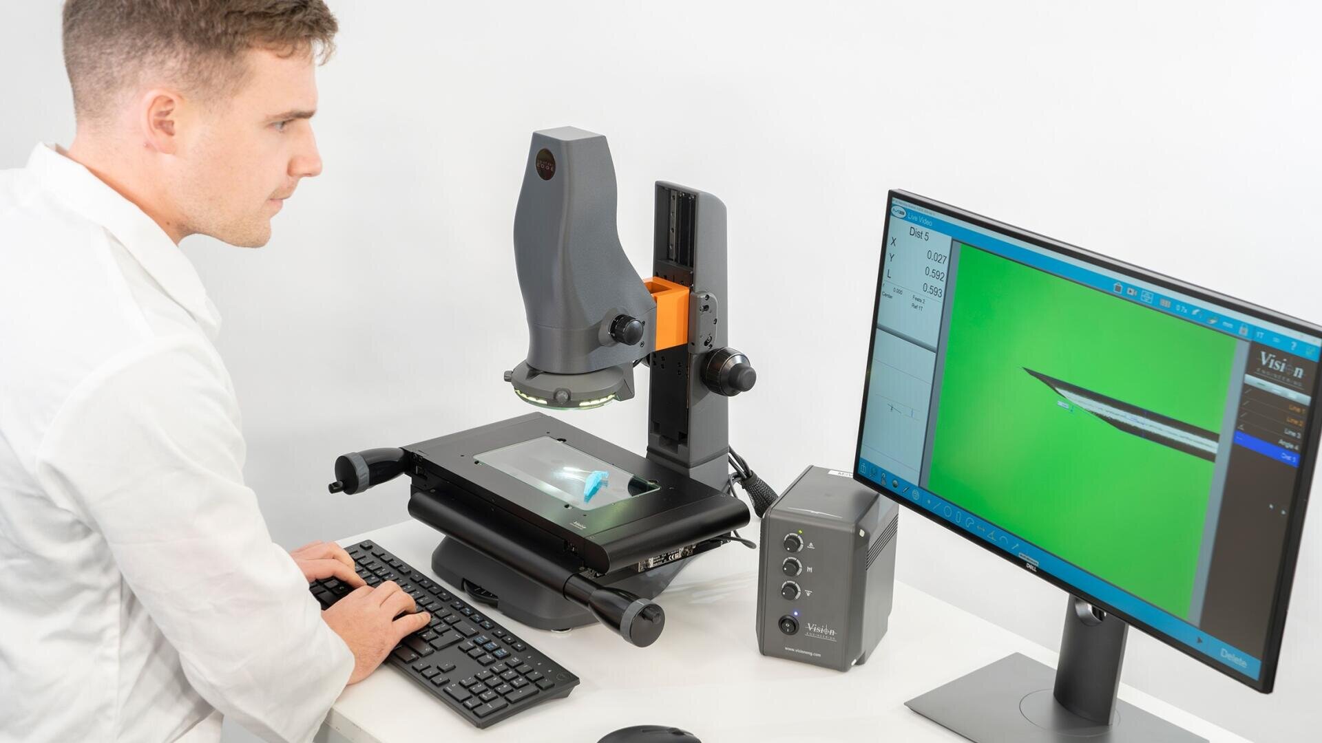 Ein Wissenschaftler in einem weißen Laborkittel untersucht eine Probe mit Hilfe eines Computers und eines digitalen Mikroskops, wobei ein Bild der Probe auf einem Monitor angezeigt wird, das eine Nahaufnahme unter Vergrößerung zeigt.