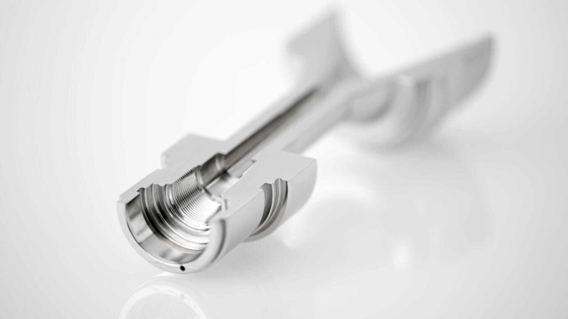 A close-up of a precision-machined metal component with internal threading and a symmetrical, cutaway design, shown on a white background.