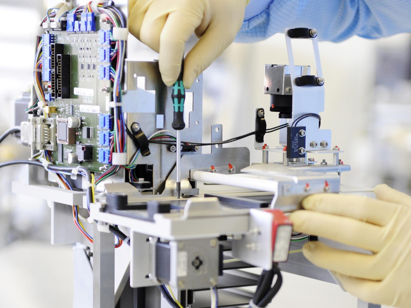 Eine Person mit Latexhandschuhen verwendet einen Schraubenzieher, um elektronische Komponenten in einer Maschine mit sichtbaren Kabeln und Leiterplatten zusammenzubauen oder zu reparieren.