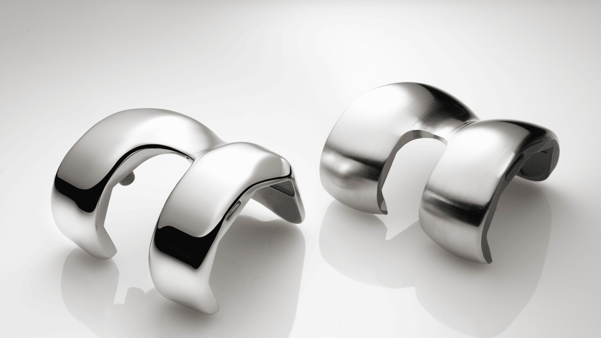 Zwei moderne, geschwungene Manschettenarmbänder aus silbernem Metall mit glatten, glänzenden Oberflächen und abstrakten, offenen Mustern liegen auf einer reflektierenden weißen Oberfläche.