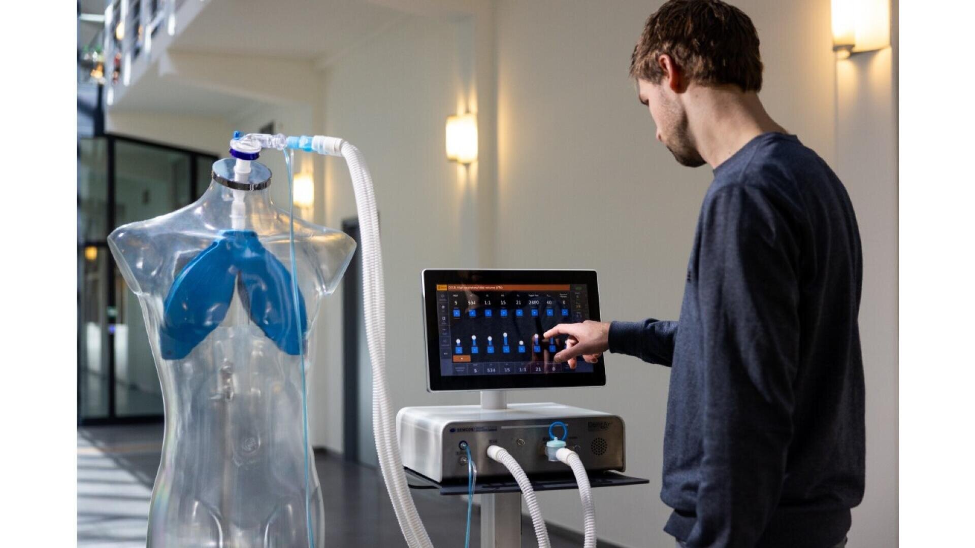 Ein Mann verwendet ein Touchscreen-Beatmungsgerät, das an einen durchsichtigen Puppentorso mit blauen Lungen angeschlossen ist. Wahrscheinlich demonstriert oder testet er medizinische Geräte in einem hellen, modernen Innenraum.