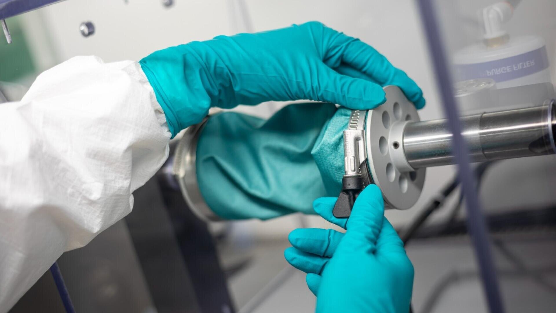 A person in a protective suit and teal gloves adjusts equipment with a metal cylindrical component inside a laboratory glove box, suggesting a controlled or sterile work environment.
