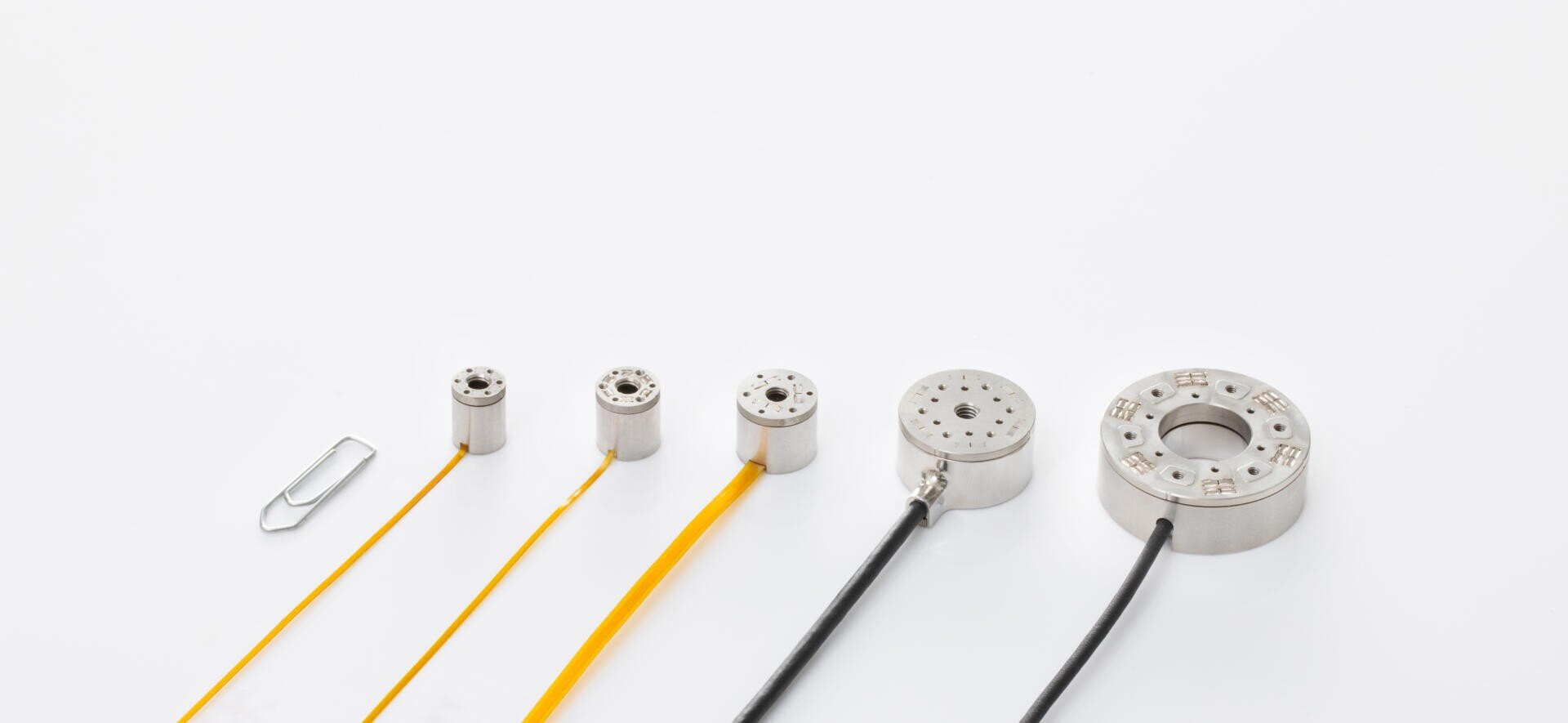 Five circular metal devices of increasing size, each with attached wires, are lined up next to a paper clip on a white background to illustrate size.
