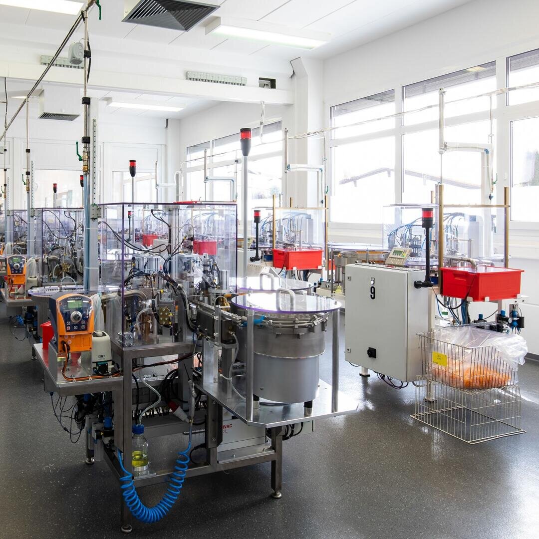 Eine helle, moderne Fabrikhalle voller automatisierter Maschinen und Anlagen. Mehrere Maschinen aus Metall und Kunststoff mit transparenten Abschirmungen stehen in Reihen auf einem glänzend dunklen Boden, große Fenster lassen Tageslicht herein.