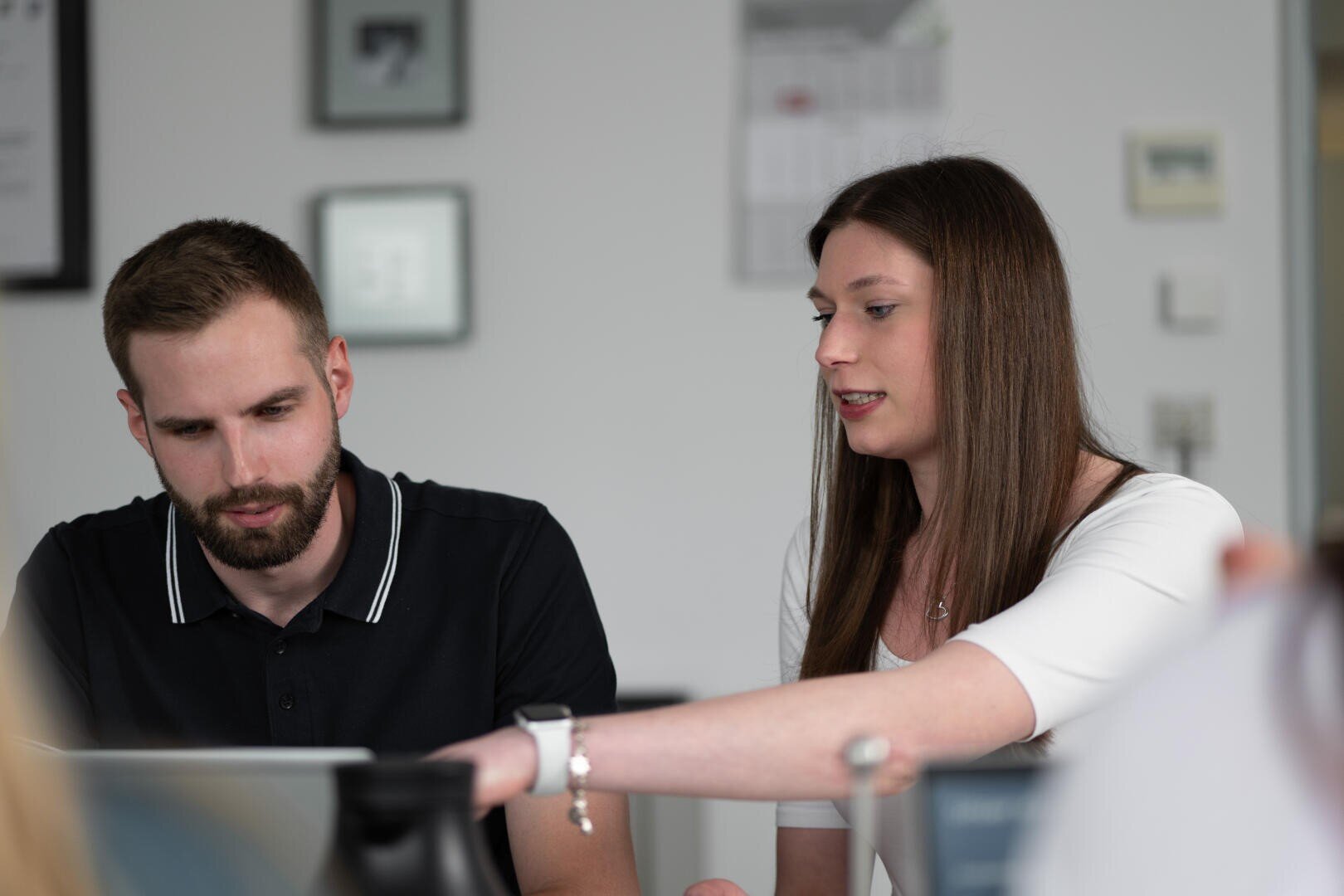 A woman points at a laptop screen while speaking to a man sitting beside her. Both appear focused and engaged in a discussion in a bright office setting.