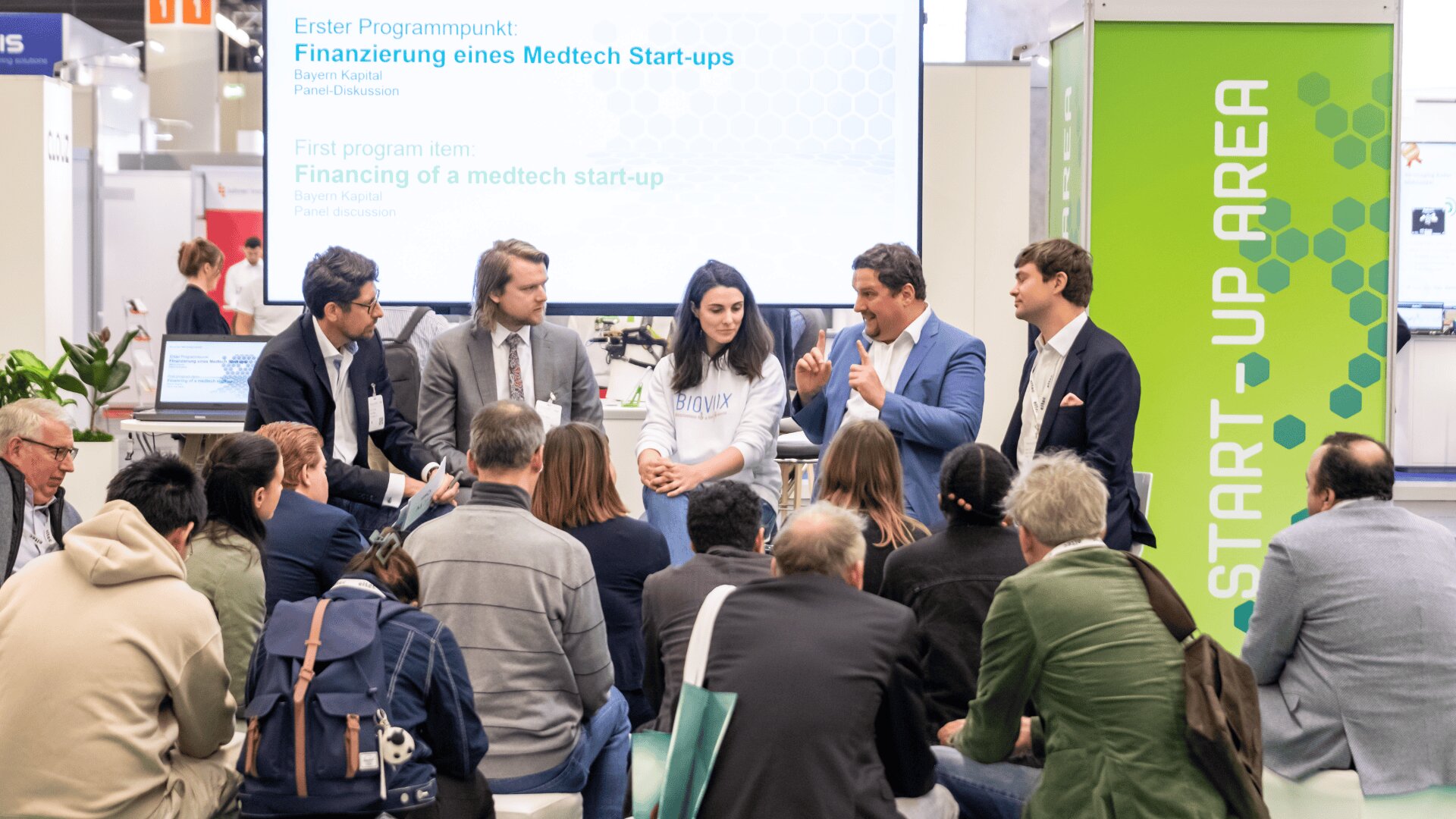 Eine Gruppe von Menschen sitzt auf einer Konferenz und hört einer Podiumsdiskussion über die Finanzierung eines Medtech-Start-ups zu. Fünf Diskussionsteilnehmer sitzen auf Stühlen und sprechen vor einem Bildschirm und einem grünen Banner mit der Aufschrift „START-UP AREA“.