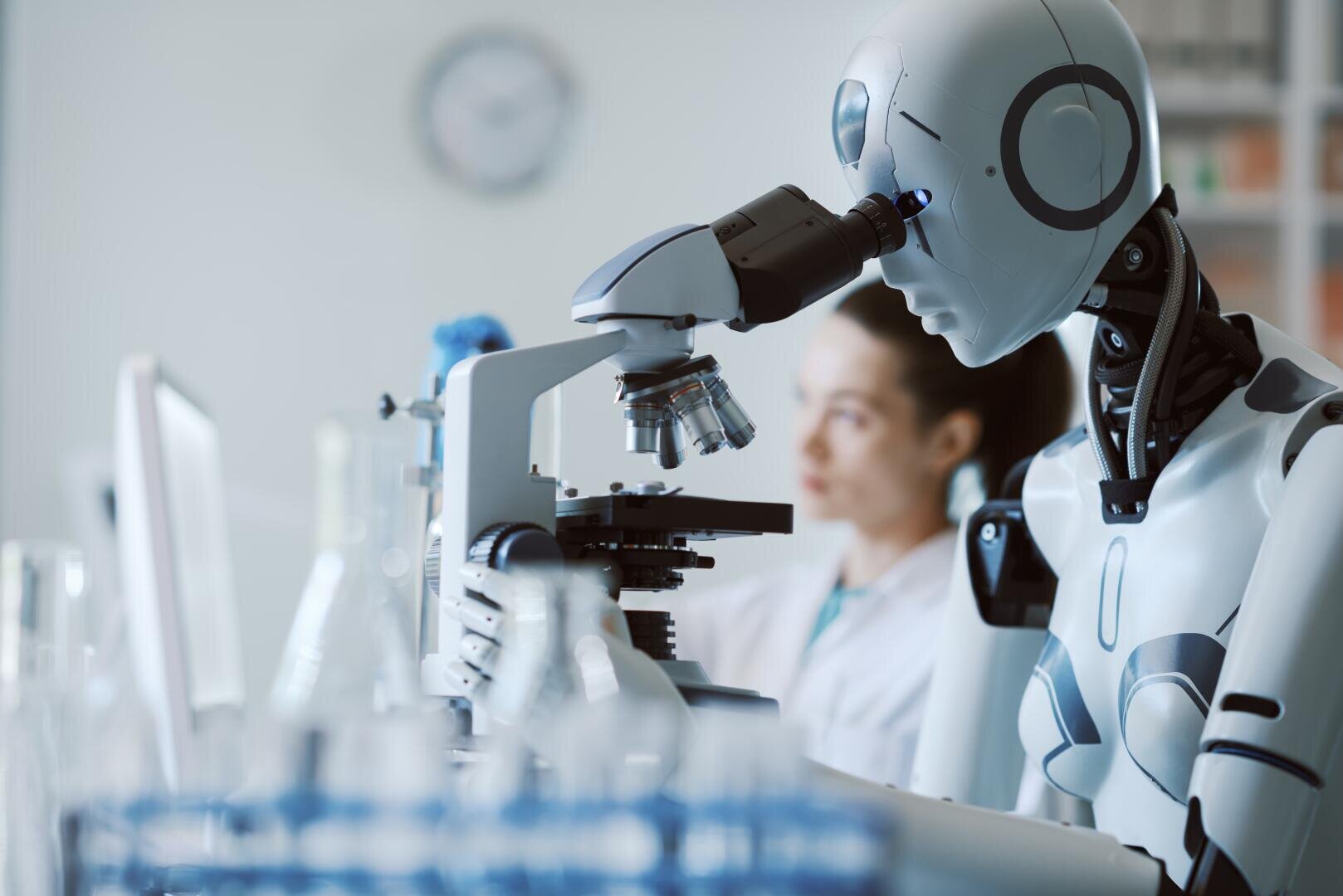 A humanoid robot examines a sample through a microscope in a laboratory. Glassware lies on the table, and a blurred human scientist can be seen in the background.