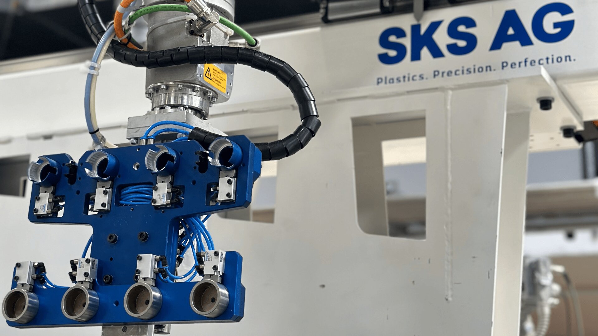 Nahaufnahme einer industriellen Roboterausrüstung mit blauen und schwarzen Komponenten, befestigt an einer Maschine mit der blauen Aufschrift „SKS AG Plastics. Präzision. Perfektion.“. Verschiedene Kabel und Anschlüsse sind sichtbar.