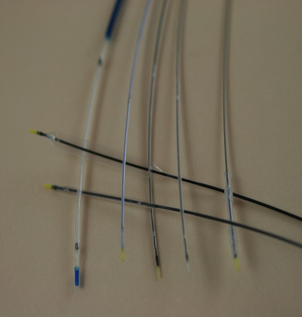 Nahaufnahme von sechs dünnen, flexiblen medizinischen Führungsdrähten auf einer beigen Oberfläche, jeweils mit farbigen Spitzen in Blau, Gelb oder Schwarz. Die Drähte sind leicht gebogen und parallel zueinander angeordnet.