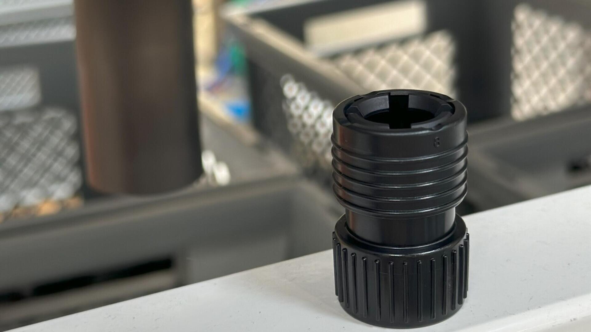 A close-up of a black plastic threaded connector or adapter resting on a white surface, with a blurred industrial or workshop background featuring metal mesh panels.