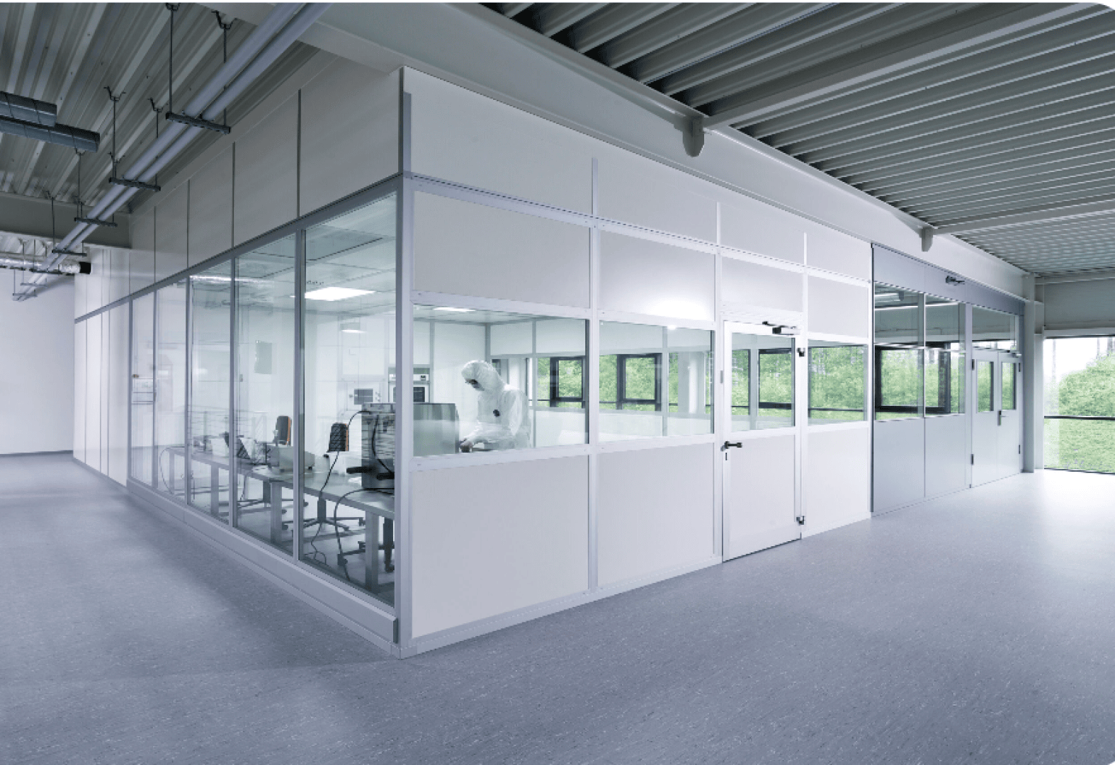 A person in protective clothing works inside a clean, modern laboratory or cleanroom with glass walls and bright lighting. The room appears sterile and organized, with equipment visible inside.