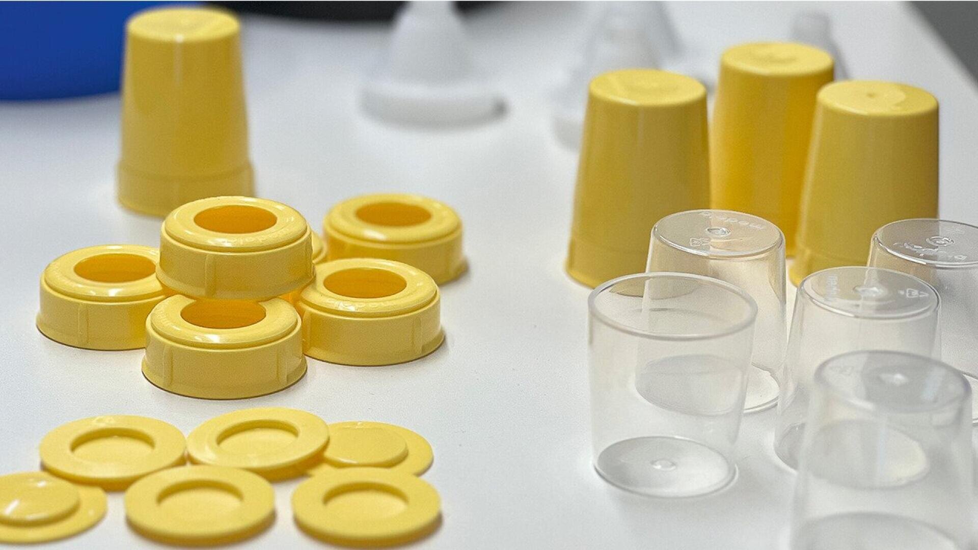 Stacks of yellow plastic bottle caps and transparent cups are arranged on a white surface, with some additional plastic pieces in the background.