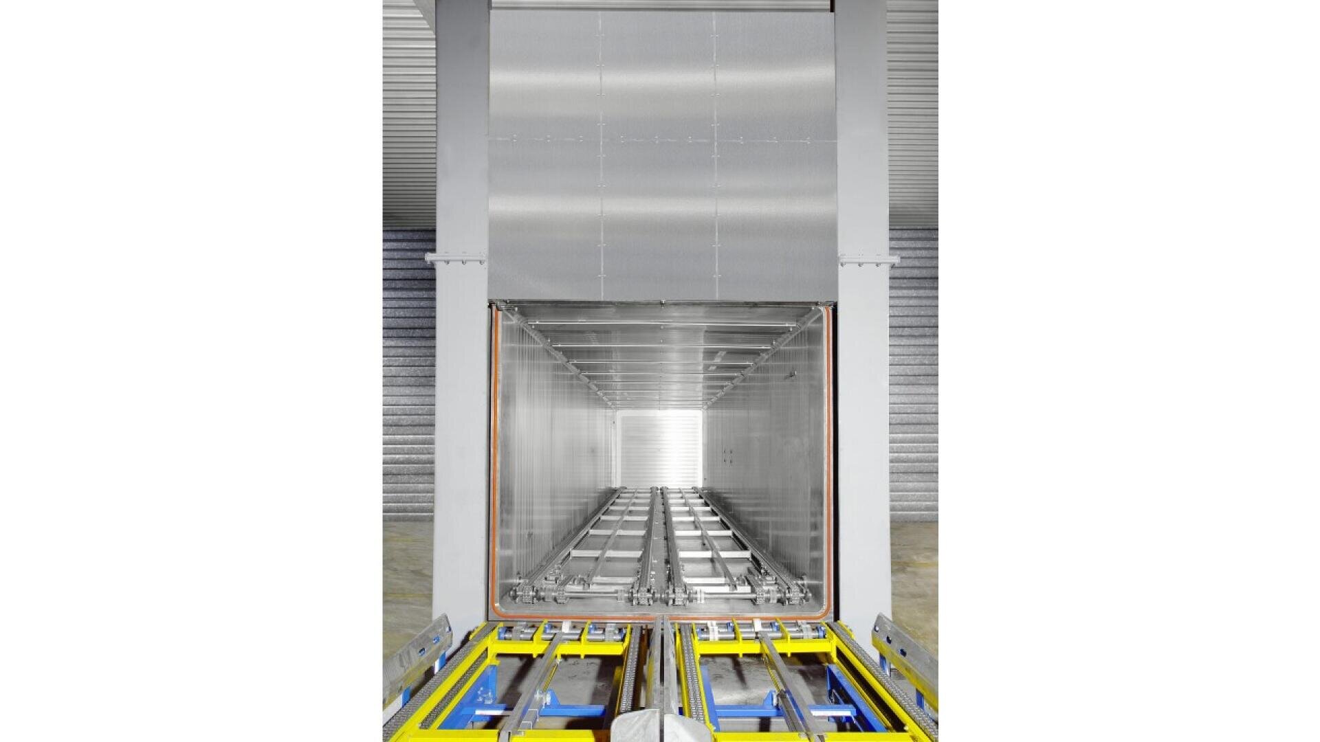 A large, empty metallic chamber with reflective surfaces and rail tracks leading inside; the view is from outside looking straight through the open entrance, with yellow and blue equipment at the front.