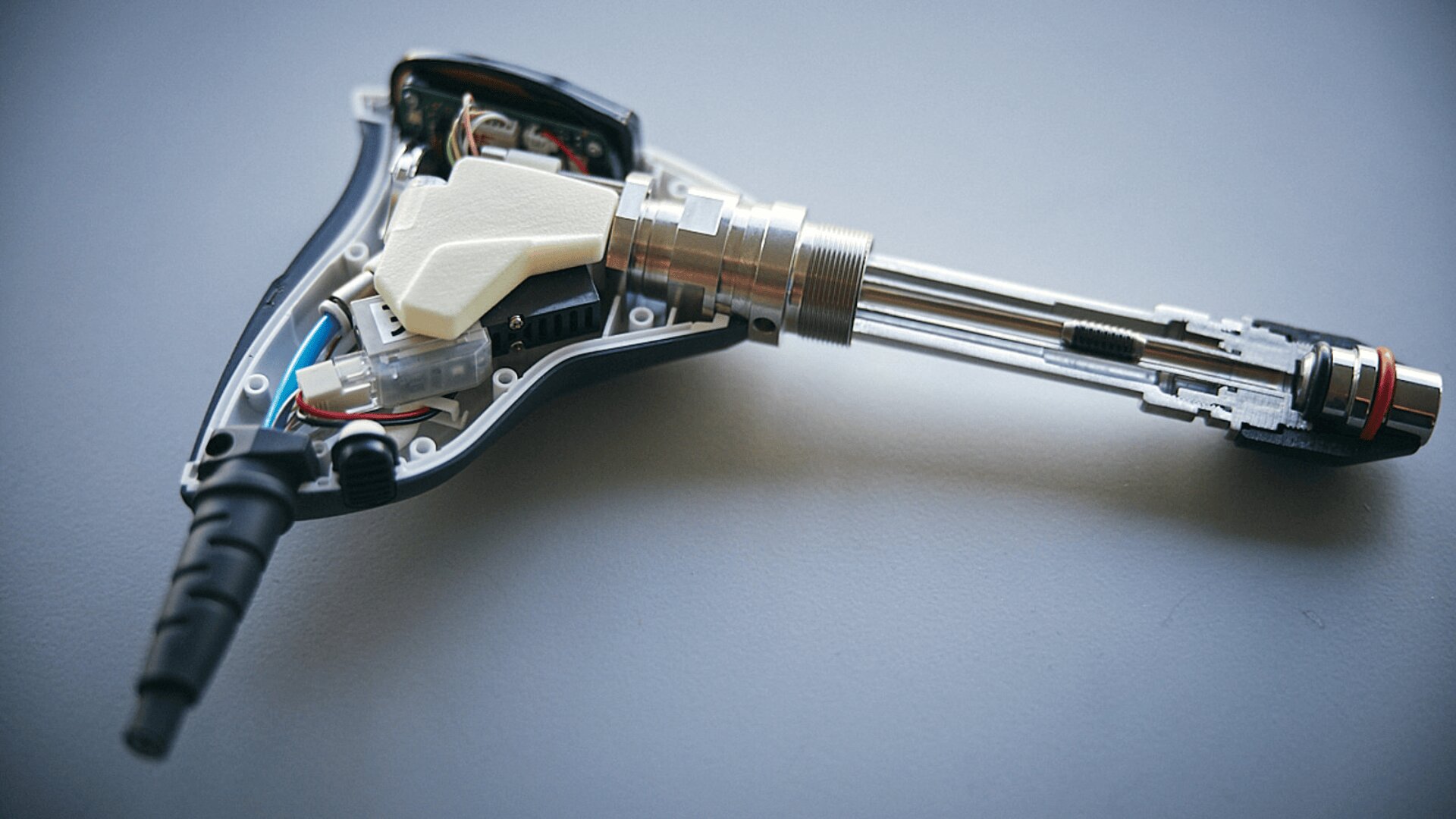 Nahaufnahme eines teilweise geöffneten elektronischen Geräts mit freiliegenden internen Komponenten, Kabeln und einem Metallschaft, dargestellt auf einer hellgrauen Fläche.