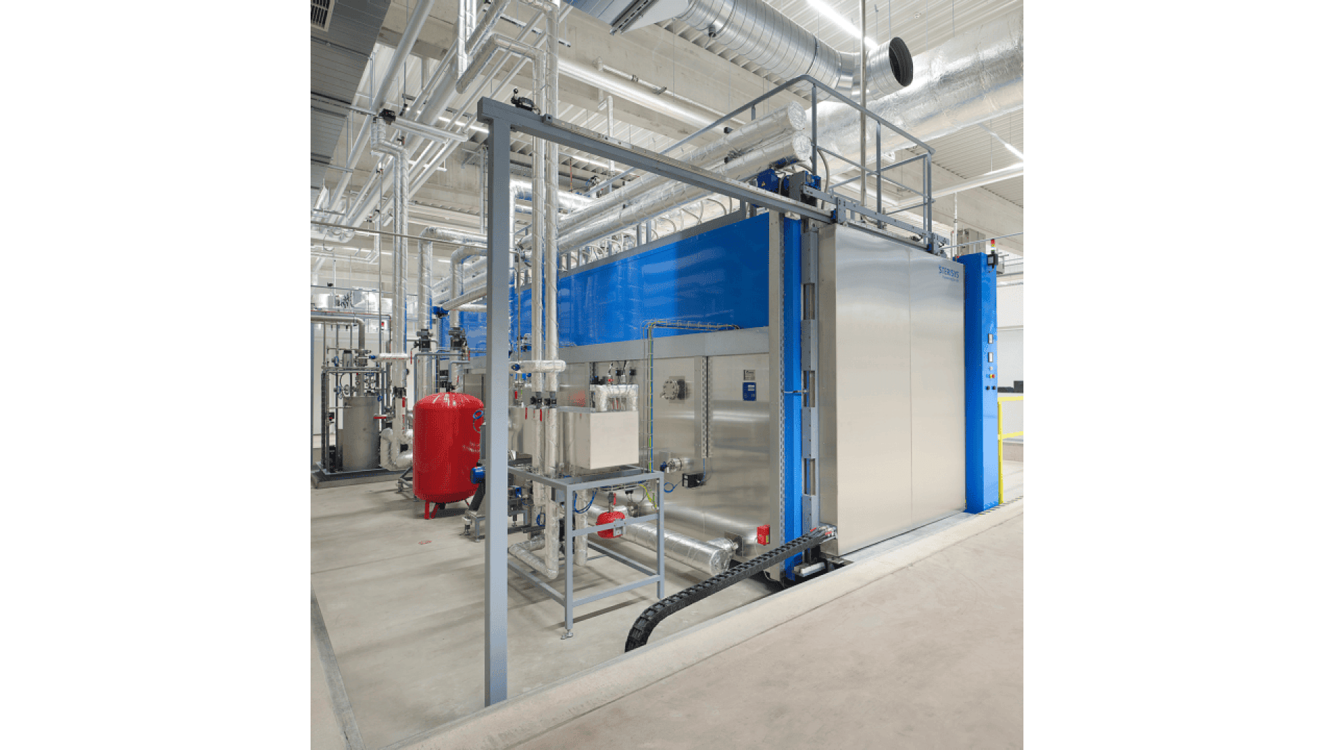 Large industrial boiler system with blue and metallic panels, surrounded by pipes, control panels, and valves, located in a clean, modern facility with visible ductwork on the ceiling.