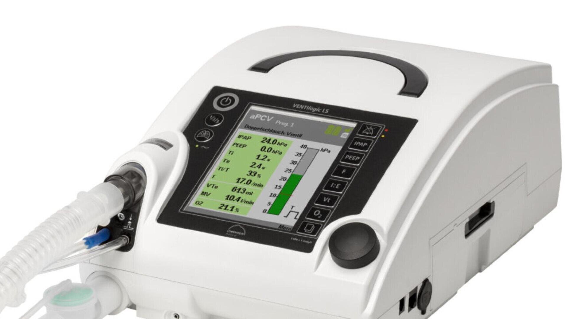 A medical ventilator with a digital display screen showing respiratory data and various settings, connected to a breathing tube. The device is white with black buttons and a handle on top.