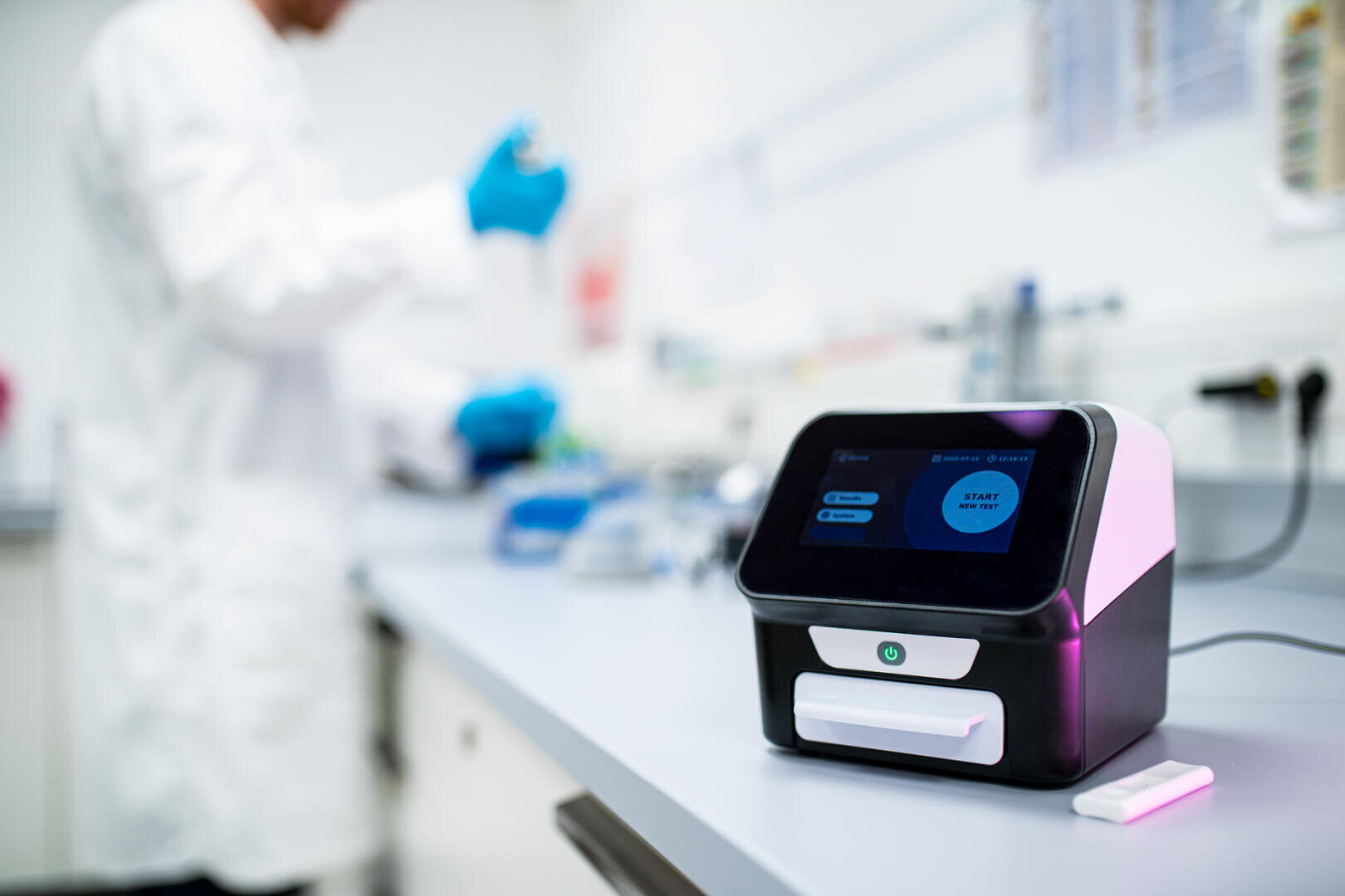 Ein Laborgerät mit Touchscreen steht auf einer Arbeitsplatte im Mittelpunkt und zeigt Optionen an. Im unscharfen Hintergrund arbeitet eine Person in einem weißen Laborkittel und blauen Handschuhen mit Laborgeräten.